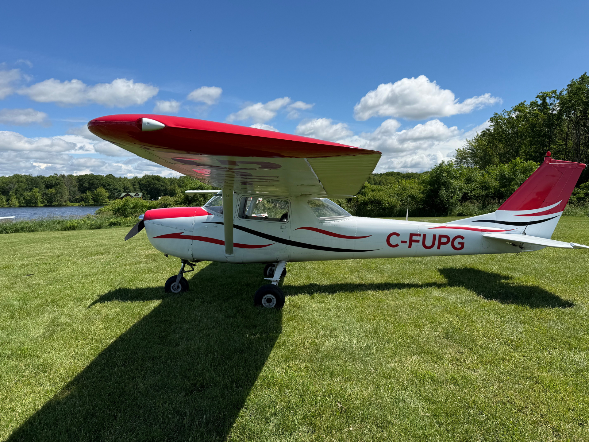 Small red-and-white single-engine airplane parked on a grassy field with blue sky and clouds; registration C-FUPG visible on fuselage.