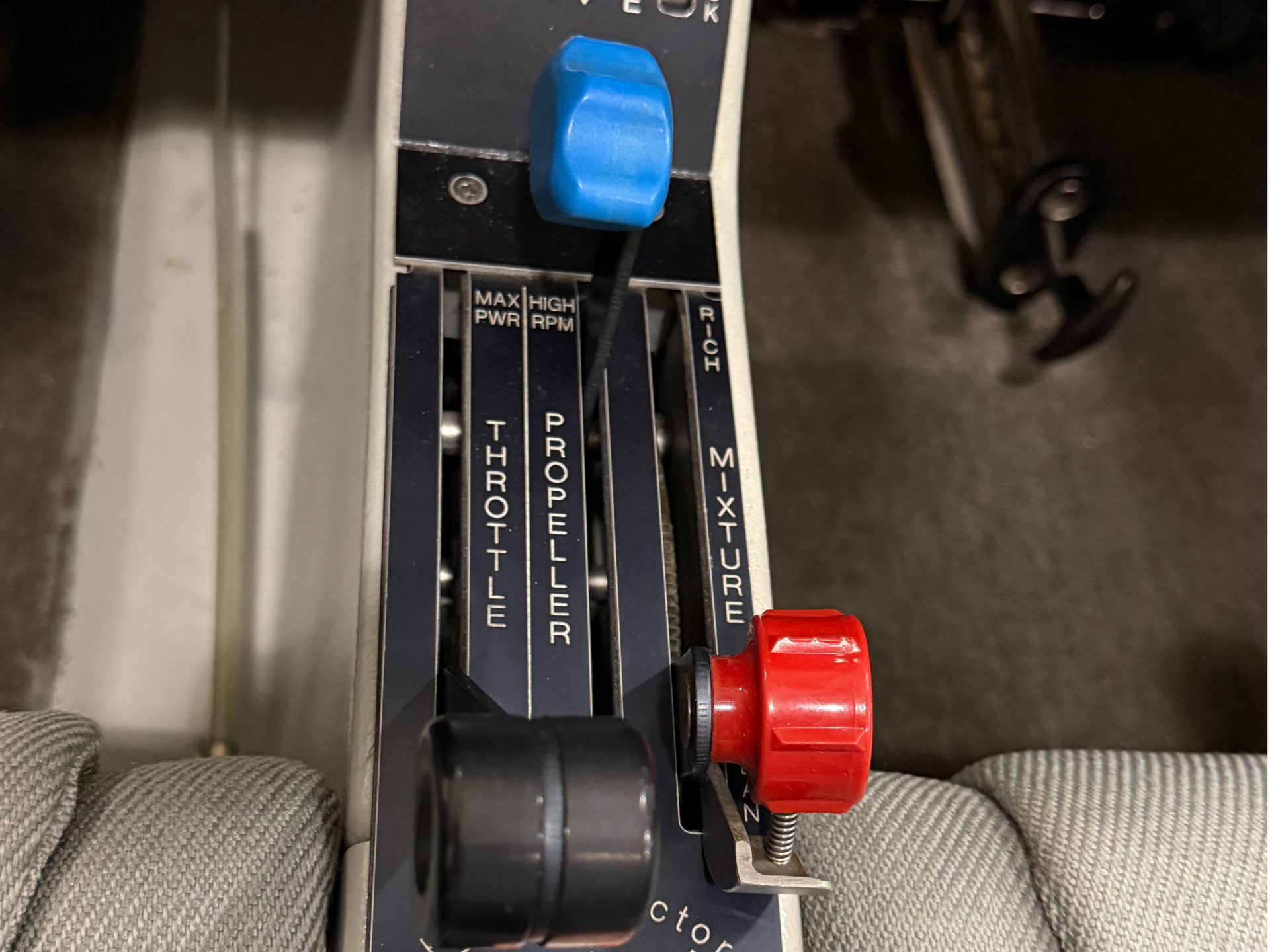 Close-up of a vintage aircraft throttle quadrant with blue control lever and red adjustment knob, levers labeled THROTTLE, PROPELLER, MIXTURE.