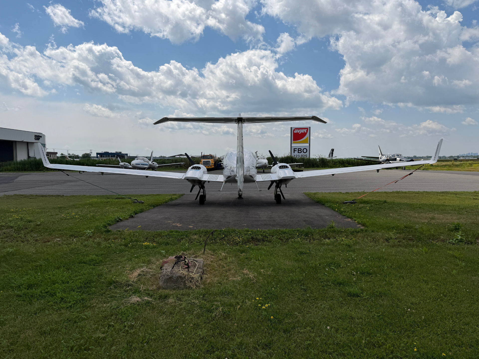 White twin-engine business jet facing the camera on a taxiway, at an airport with a hangar and fueling sign in the background.