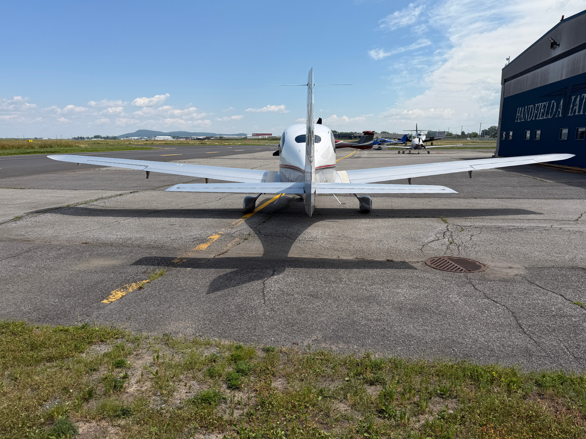 Small white single-engine airplane parked on a taxiway with a hangar on the right under a blue sky, seen from the rear cockpit.