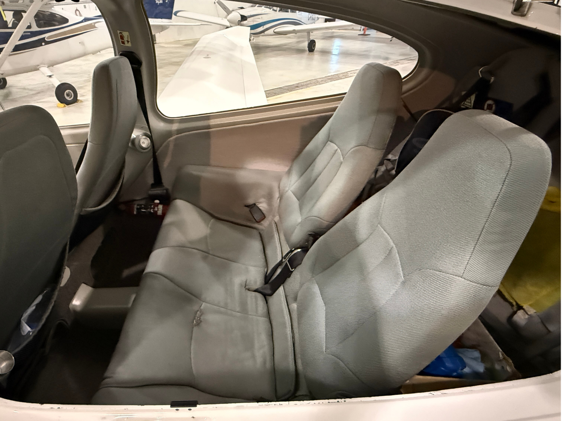 Interior of a small airplane cabin with gray fabric seats and buckled seatbelts, window showing other planes in a hangar outside.