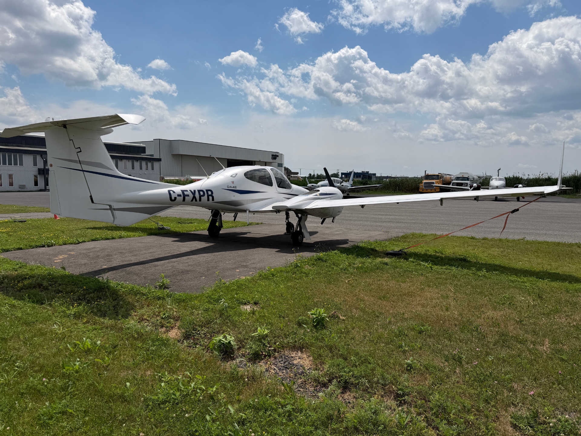 Small white Diamond DA42 aircraft parked on a taxiway with hangars in the background under a partly cloudy blue sky. Tail number C-FXPR visible.
