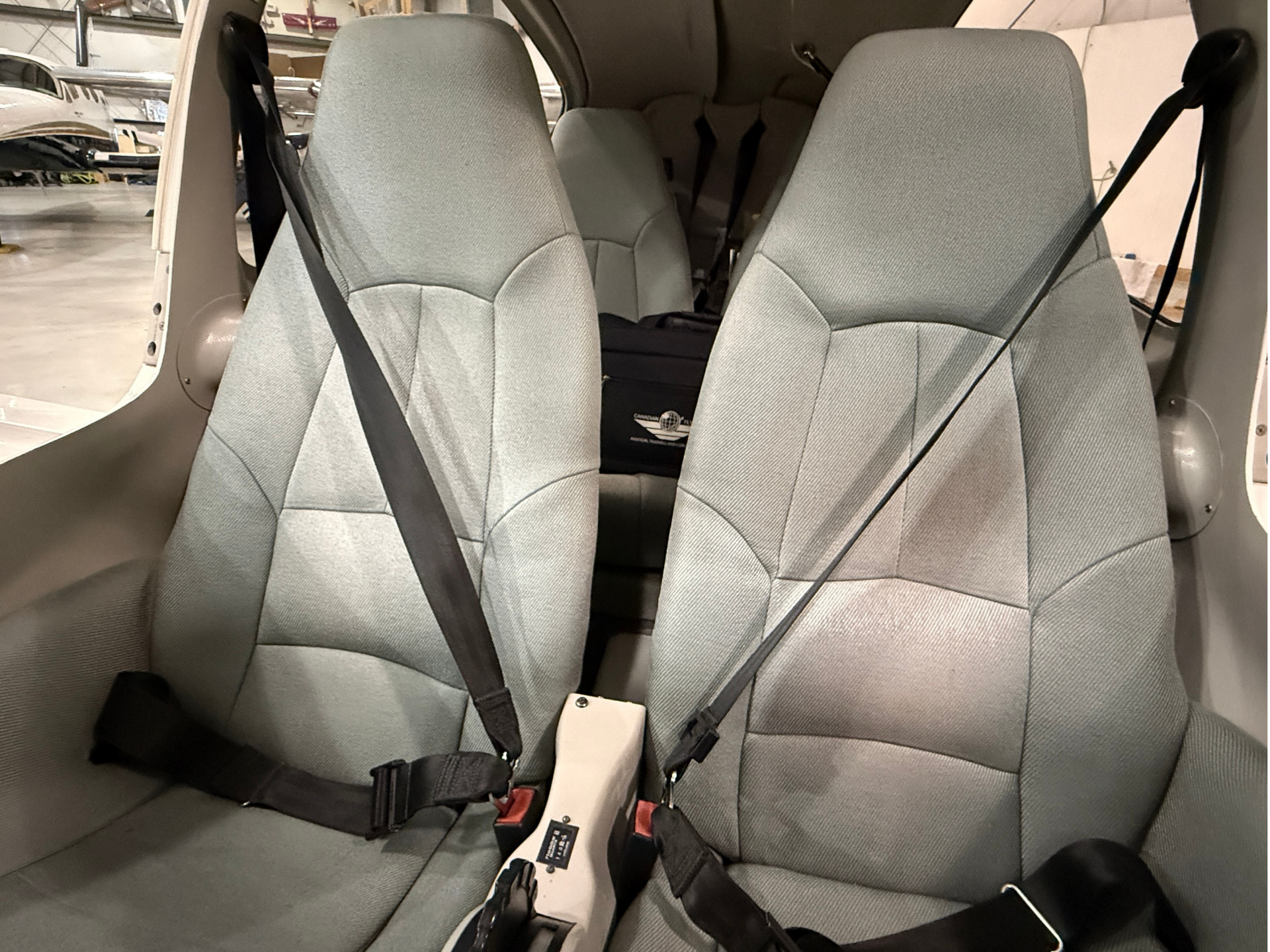 Interior of a small aircraft cabin showing gray fabric seats with overlapping lap and shoulder seat belts, cockpit area visible in background