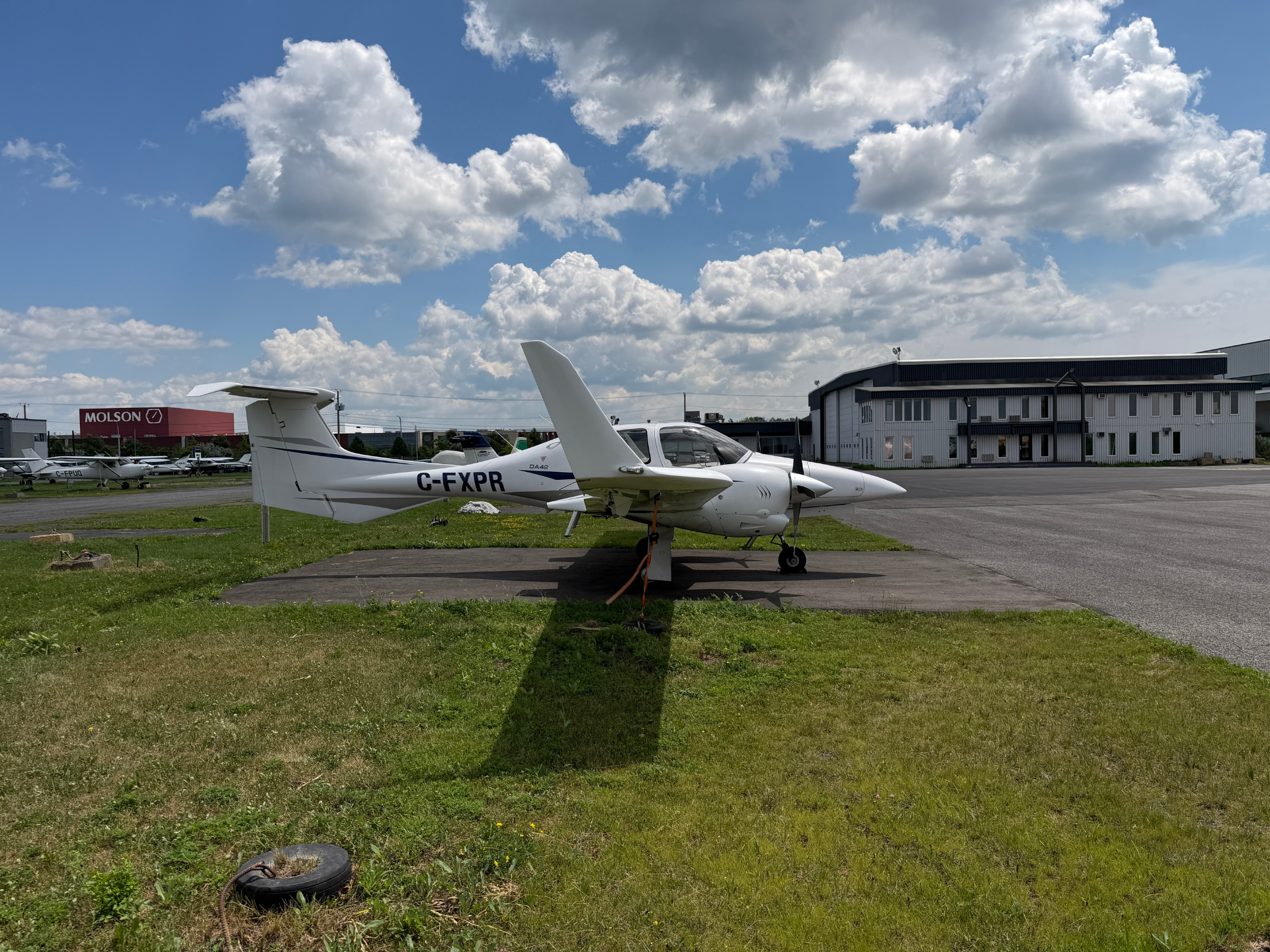 White single-engine airplane parked on a tarmac beside a hangar under a blue sky with scattered clouds.