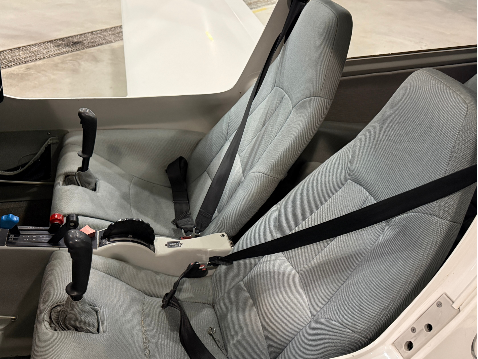 Cockpit with two gray fabric seats, seat belts, and flight yoke controls.