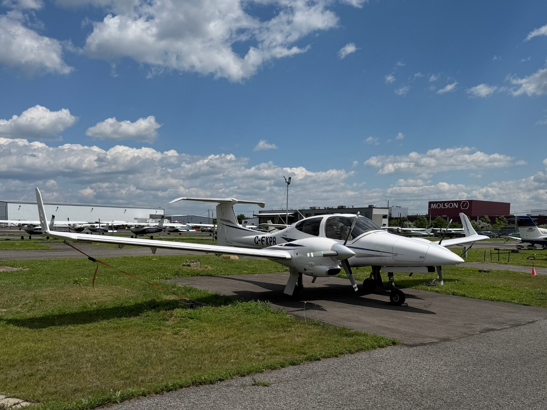Small white single-engine airplane in the foreground at an airport, with more planes, hangars, and a blue, cloudy sky in the background (registration G-FXR).