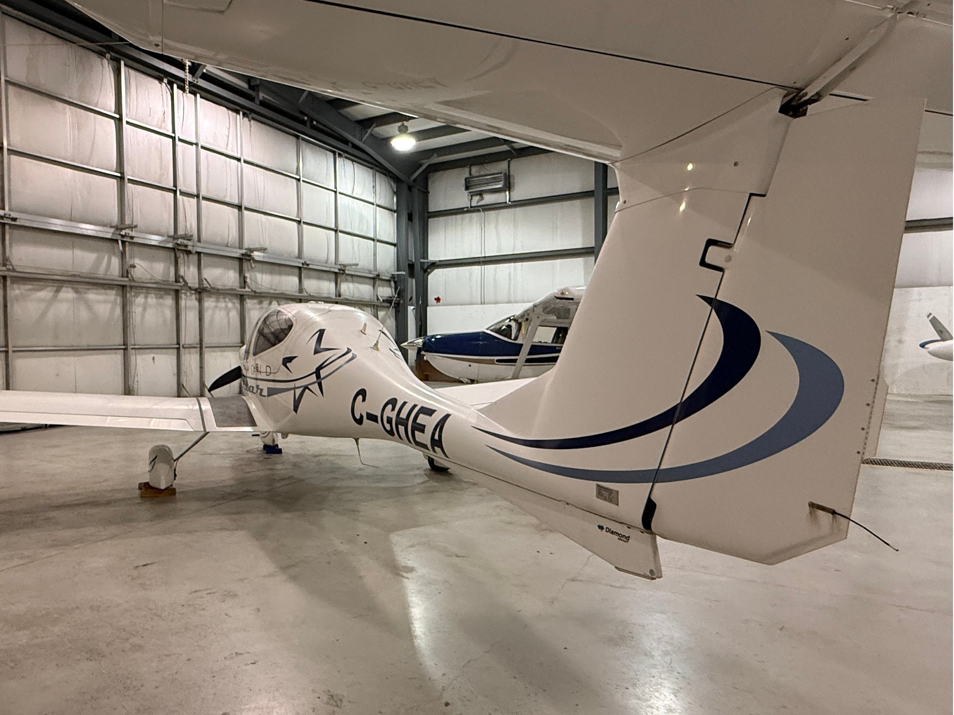 Small white airplane in a hangar, registration C-GHEA, with blue swoosh on the tail.