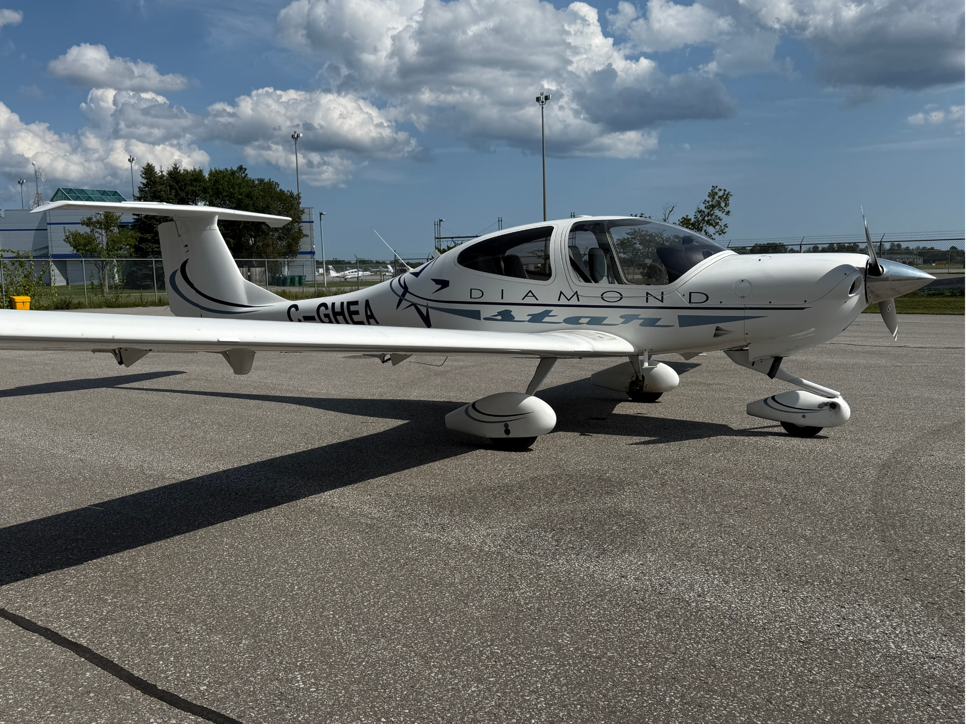 Small white Diamond aircraft parked on tarmac, propeller at nose, clear sky with fluffy clouds in background