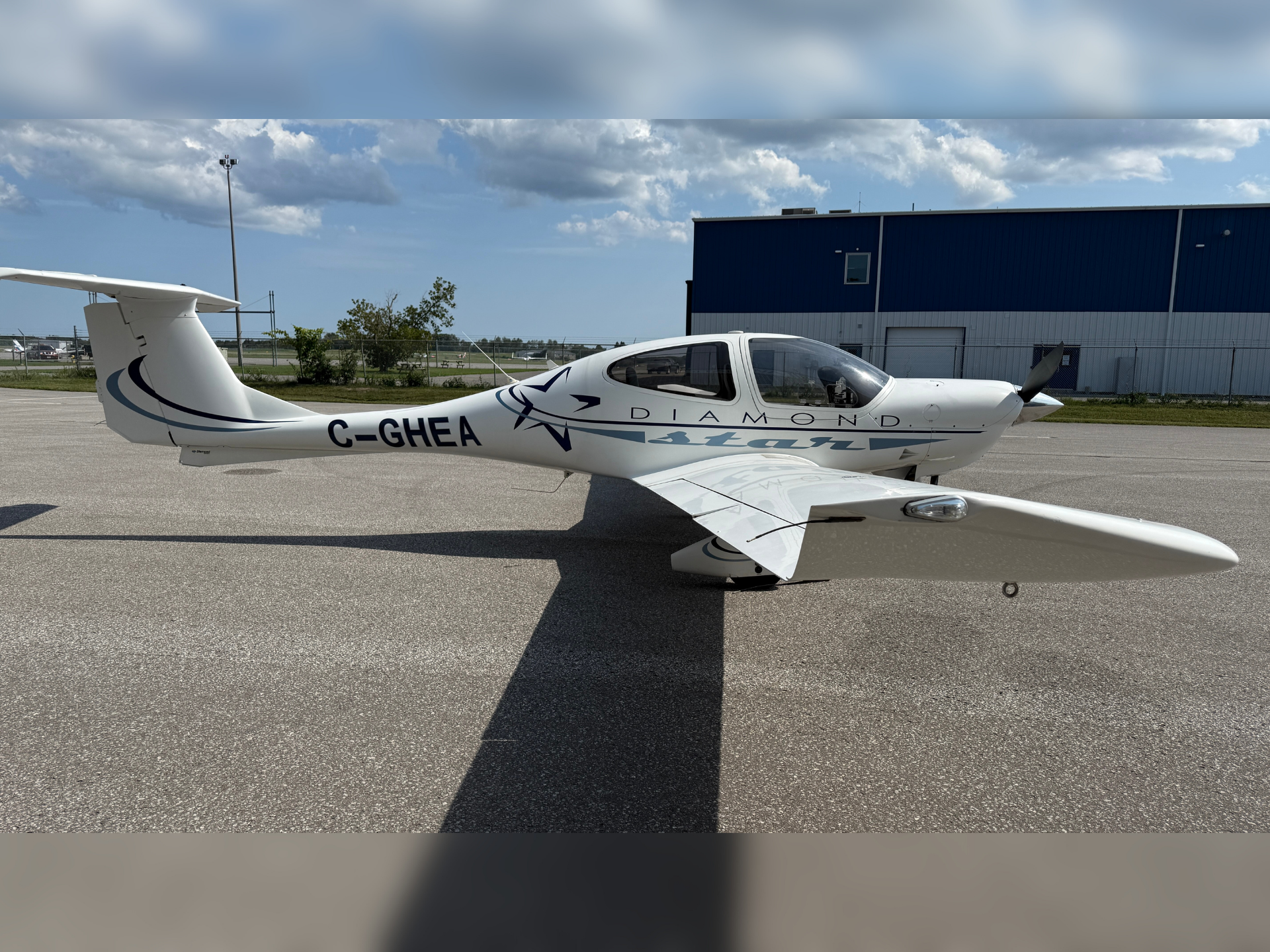Small white Diamond aircraft with registration C-GHEA parked on a tarmac near a blue hangar under partly cloudy sky.