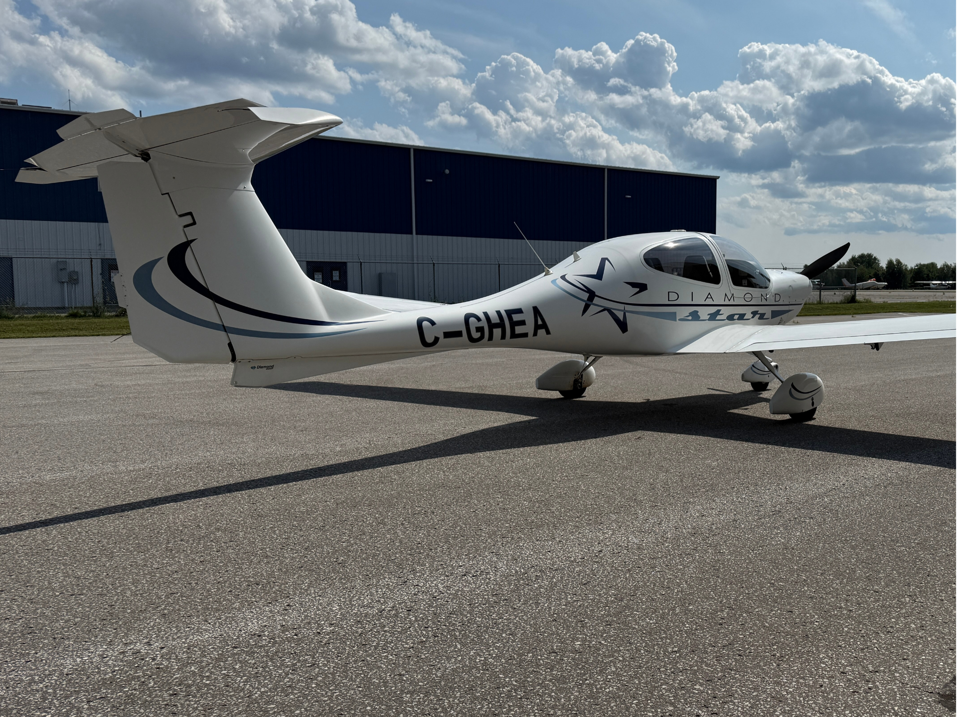 White Diamond Star airplane parked on a sunlit tarmac with a blue hangar in the background and puffy clouds overhead, registration C-GHEA visible on the fuselage.