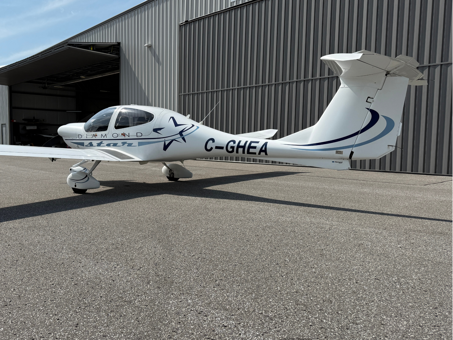 Small white Diamond light aircraft with registration C-GHEA parked on the tarmac beside an open hangar and blue decorative graphics on the fuselage.