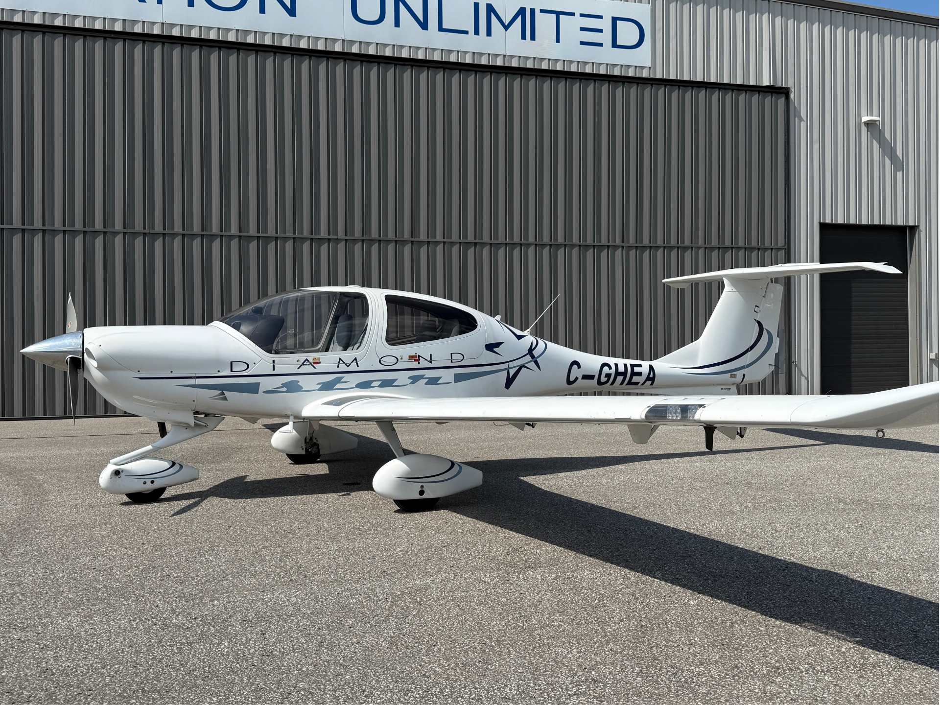 White single-engine propeller airplane parked on tarmac in front of a gray metal hangar; 'DIAMOND' branding on the fuselage and C-GHEA visible on the tail area