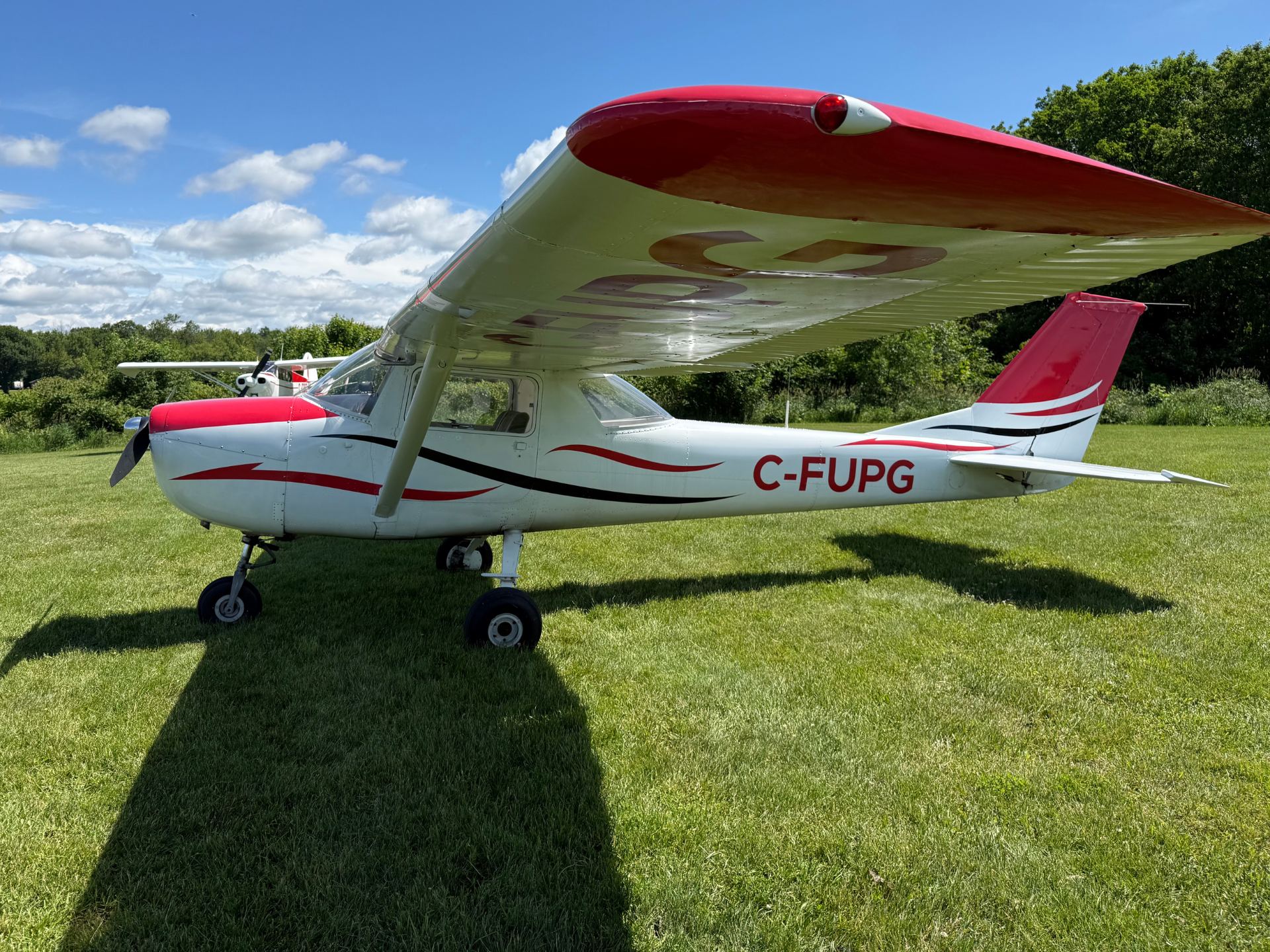 Small white and red propeller plane parked on a grassy airfield under a blue sky with scattered clouds on a sunny day.