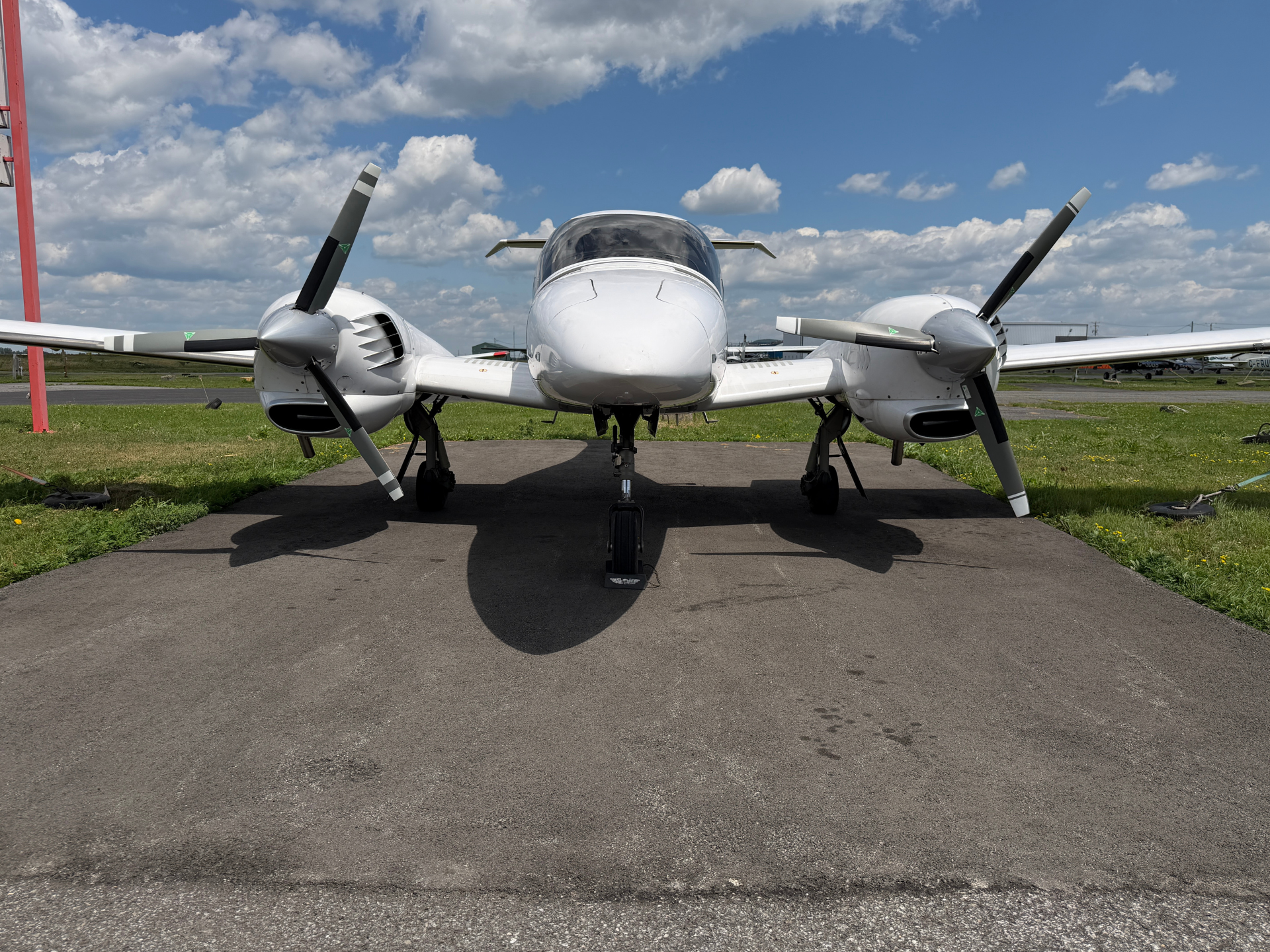 Front view of a white twin‑engine propeller airplane parked on a tarmac under a blue sky with scattered clouds on a sunny day to the side grass area visible in background]