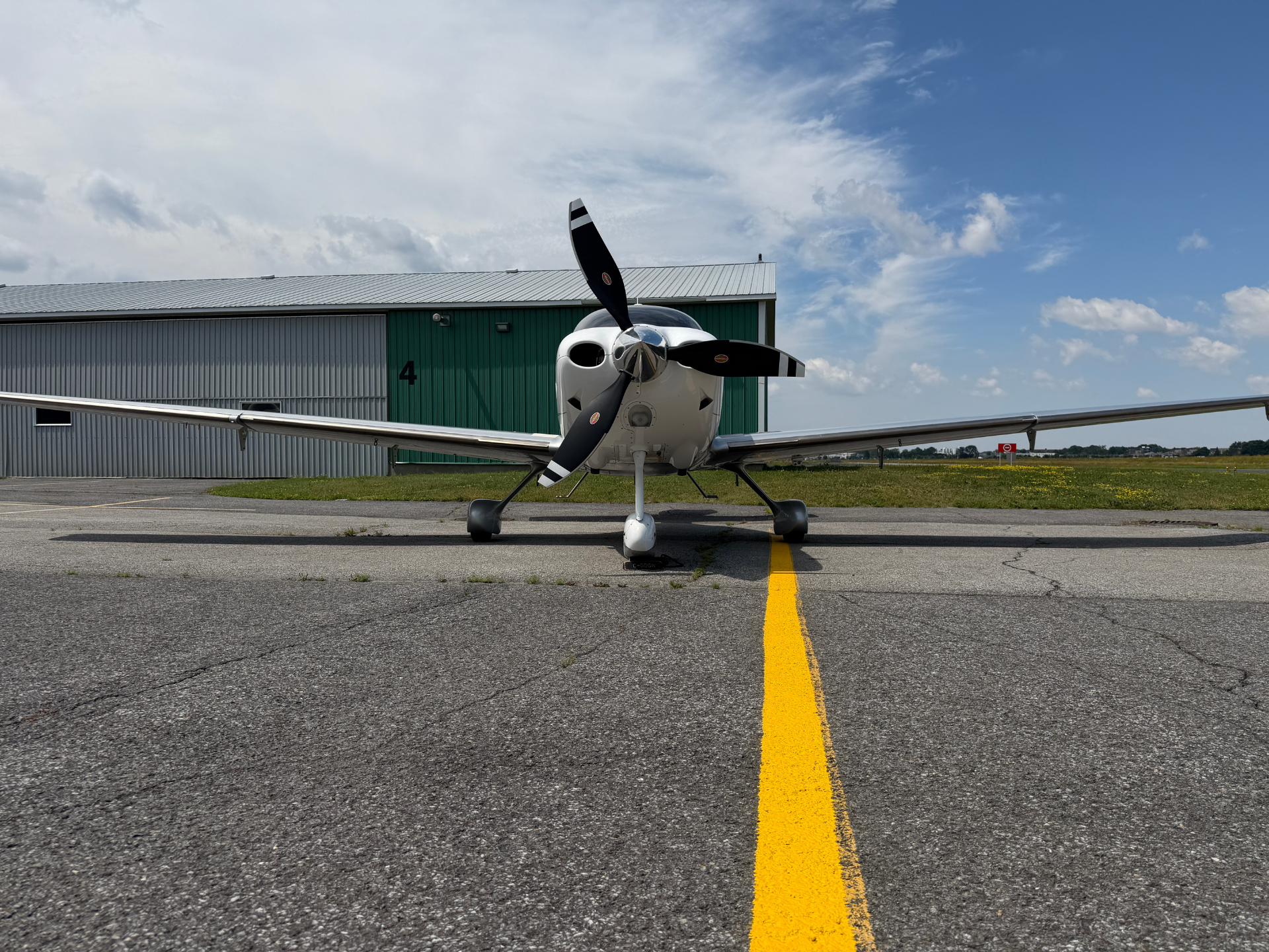 Front view of a small twin-prop airplane parked on a runway with a yellow centerline and a hangar in the background.