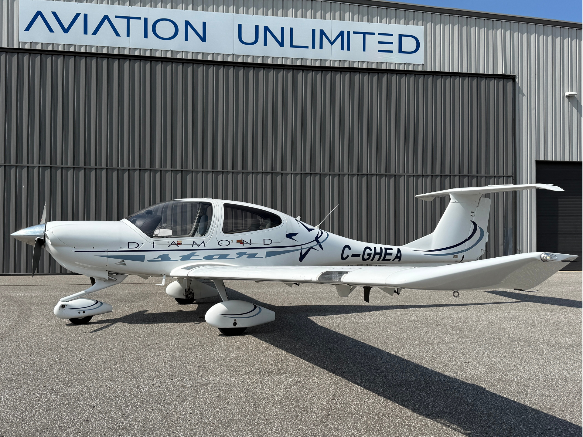 White single-engine Diamond aircraft parked on tarmac in front of a corrugated hangar with 'AVIATION UNLIMITED' sign above the doors, registration C-GHEA visible