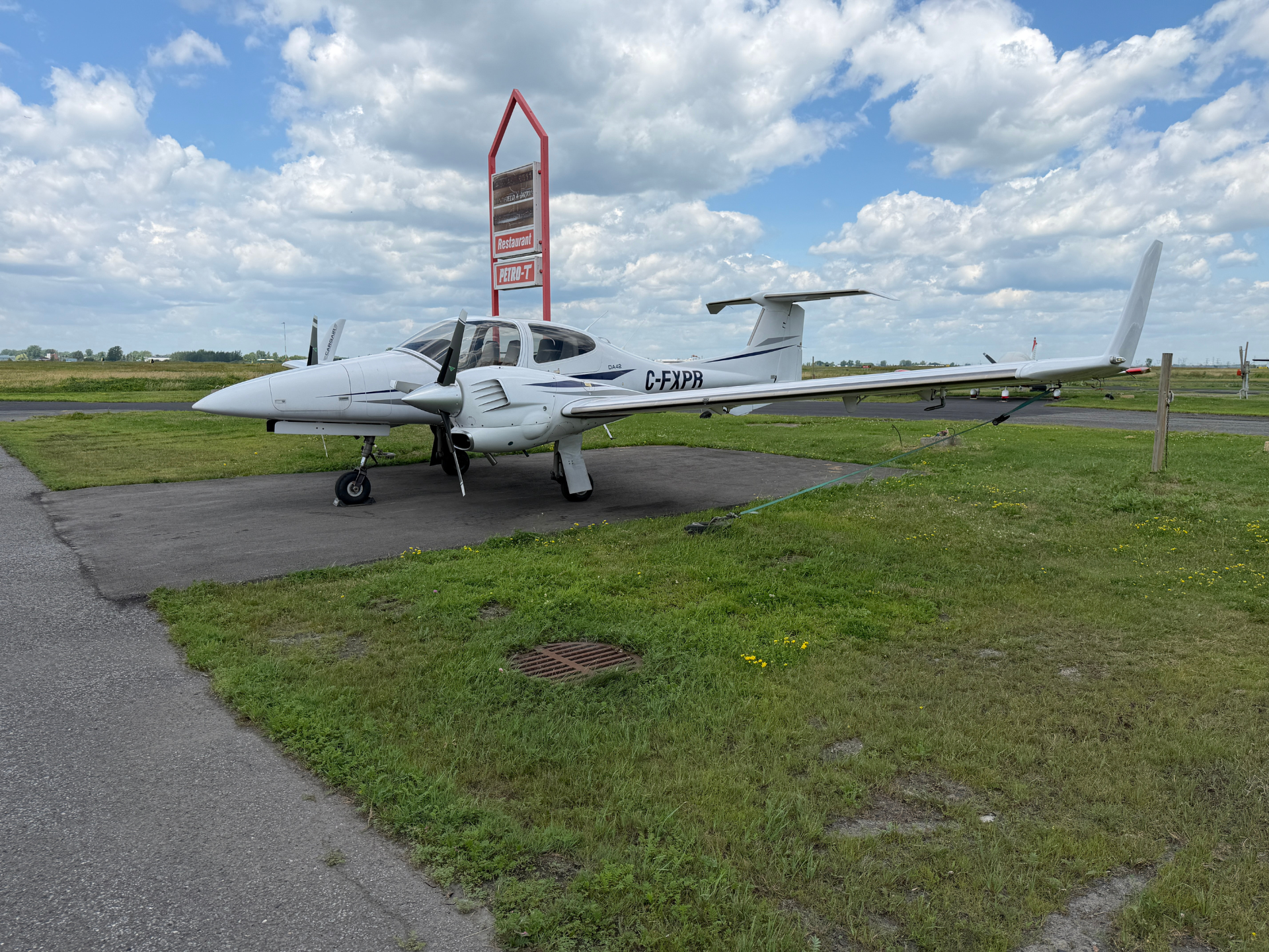White twin-engine airplane parked on a grassy airfield beside a red-and-white sign and a clear, cloudy sky above.