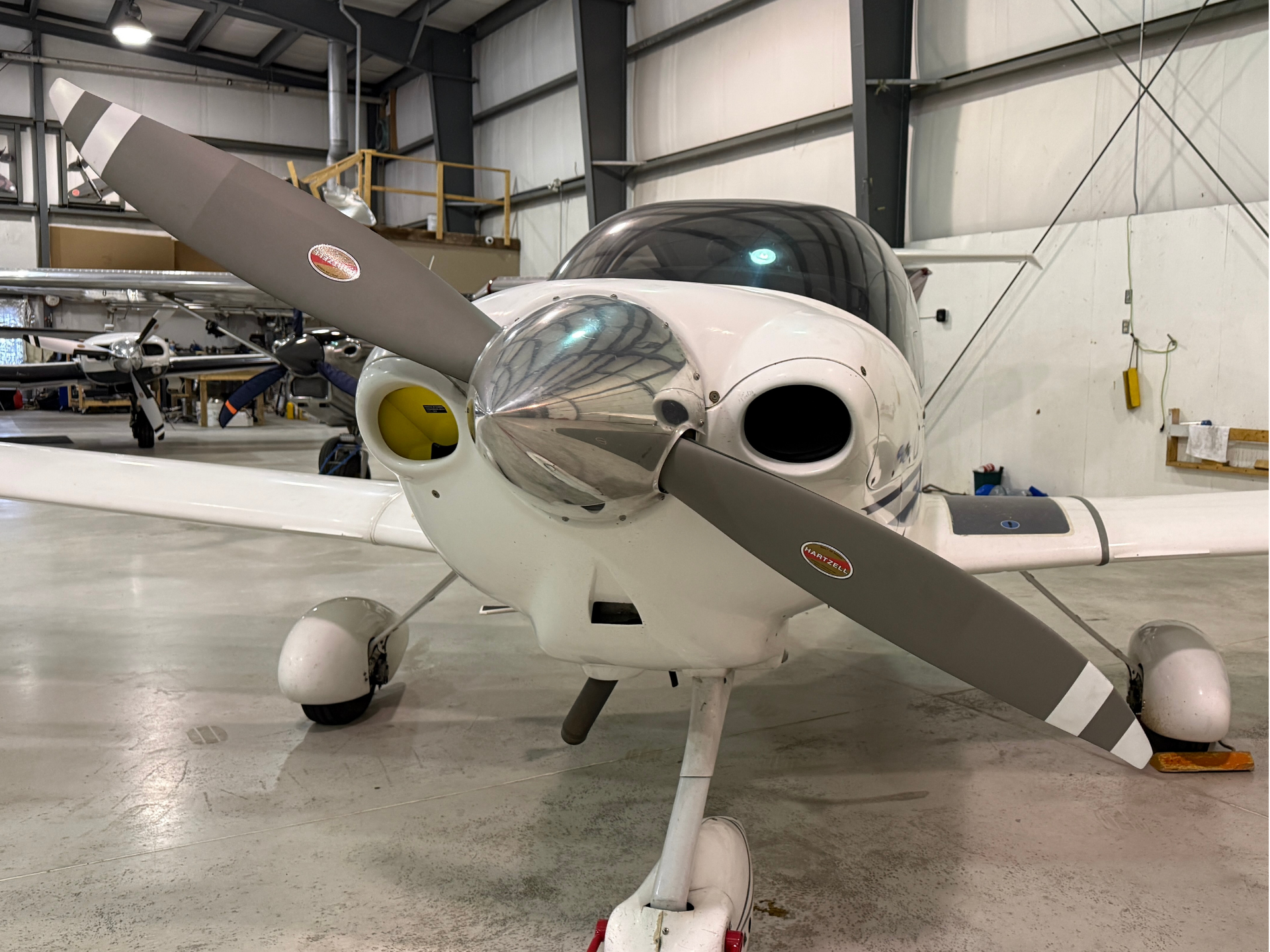 Small single-engine airplane inside a hangar, close-up of the nose and propeller with Hartzell logo visible