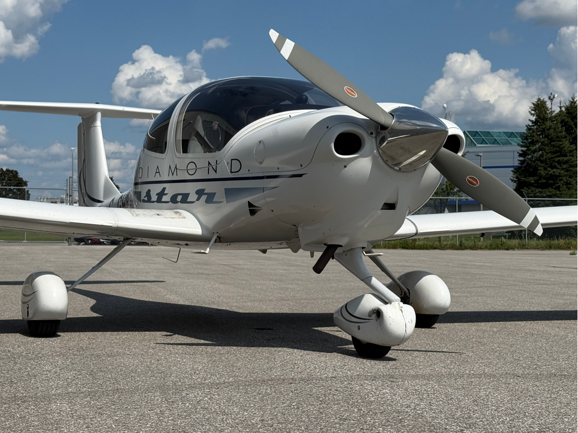 Front-left view of a small white Diamond aircraft on a tarmac, featuring a two-blade propeller and tricycle landing gear under a blue sky.