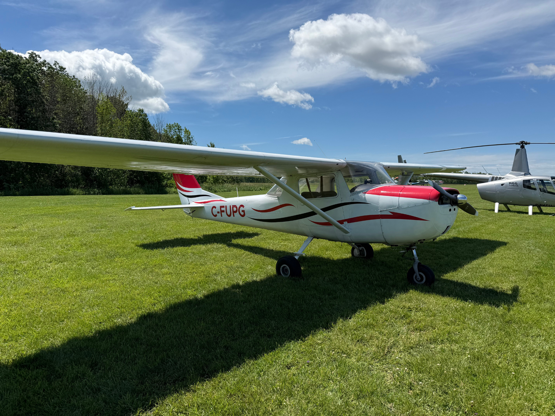 Small white and red light aircraft on a grassy airfield with a blue sky and a distant helicopter in the background, tail number C-FUPG visible.