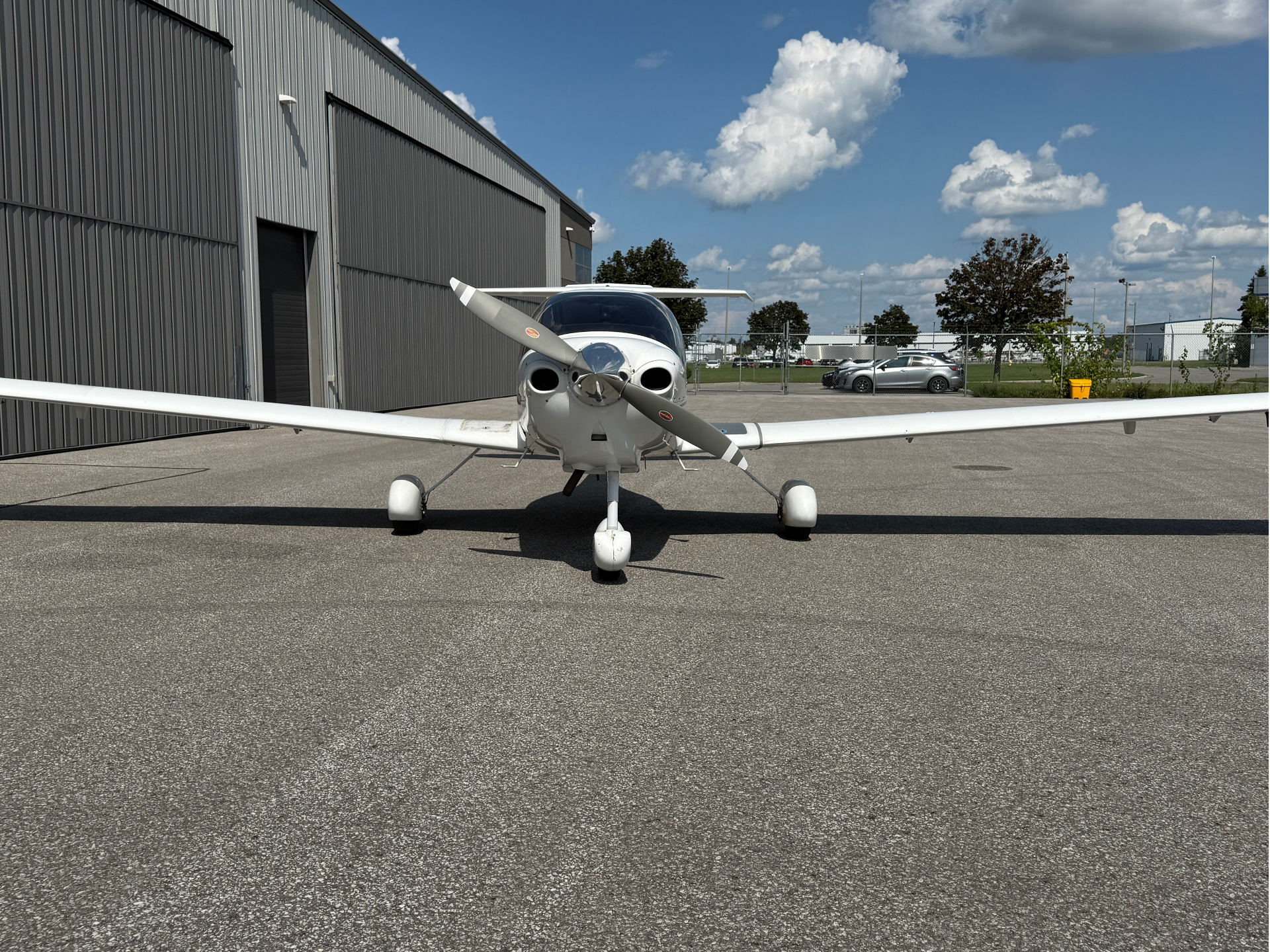 Front view of a small white single-engine airplane parked on tarmac beside a metal hangar, blue sky above.