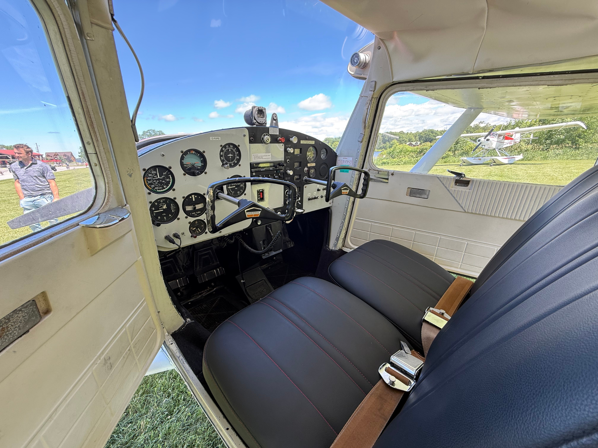 Cockpit of a small airplane with an instrument panel, yoke, and seat belts; outside shows a bright blue sky and a parked plane on the grass outside the window.