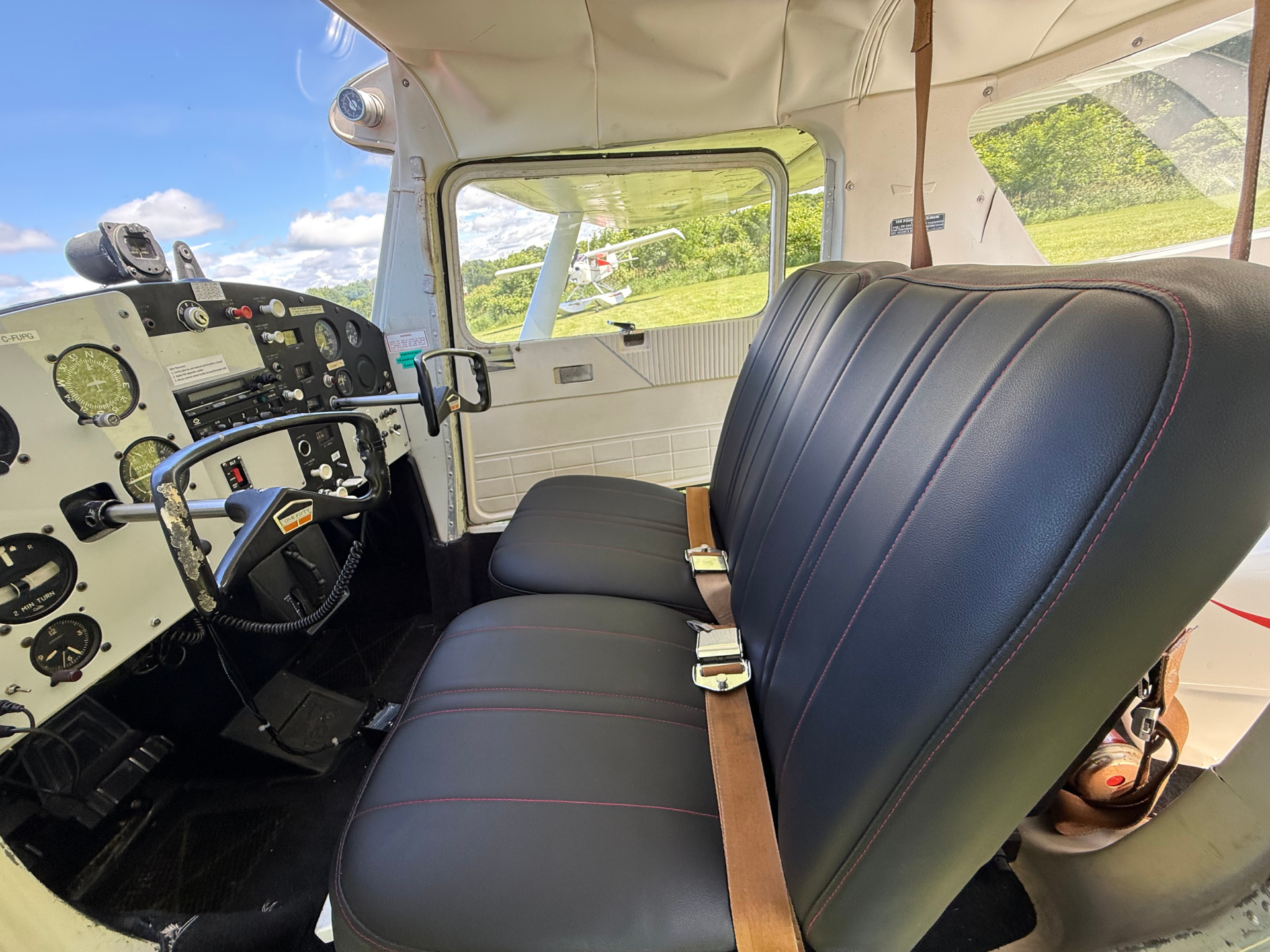 Cockpit of a small airplane with an analog instrument panel and control yoke, a blue leather seat with a brown seat belt, and a view of a grassy airfield with another plane outside through the window.