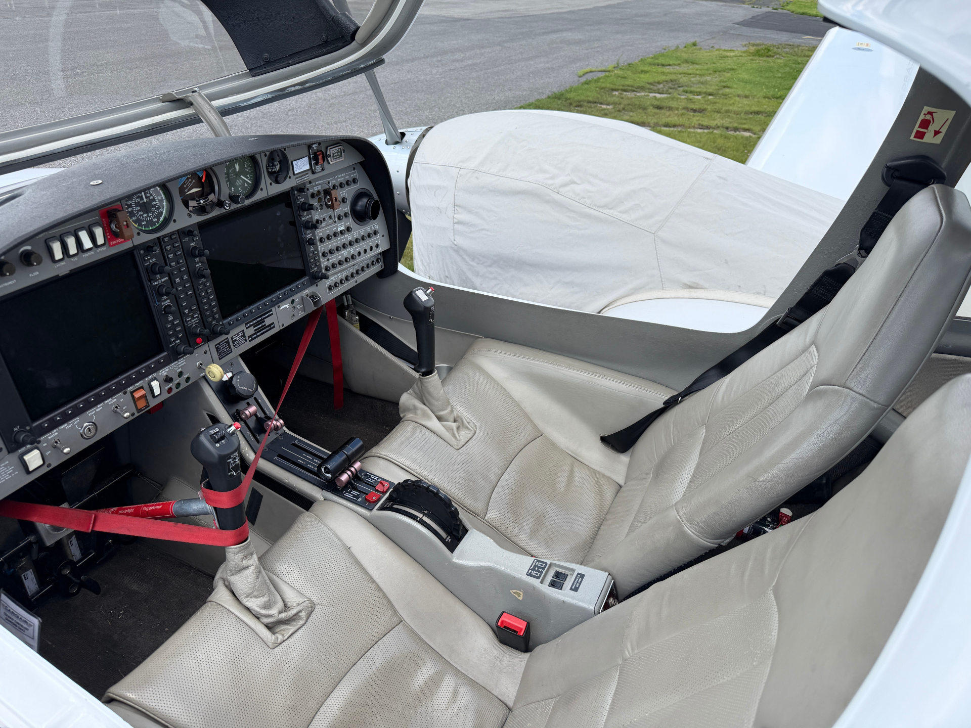 Cockpit of a small airplane with analog gauges, joysticks, and beige seats; canopy open and runway visible outside