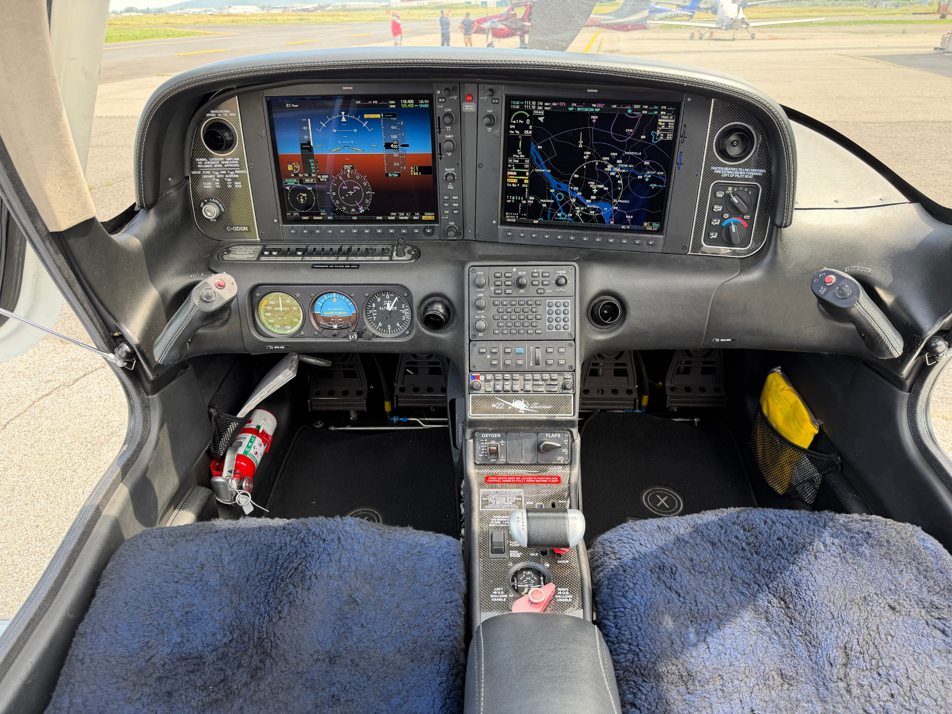 Portrait view of a small aircraft cockpit with two large instrument displays, center console, and controls, viewed from the pilot seat at the airstrip edge.
