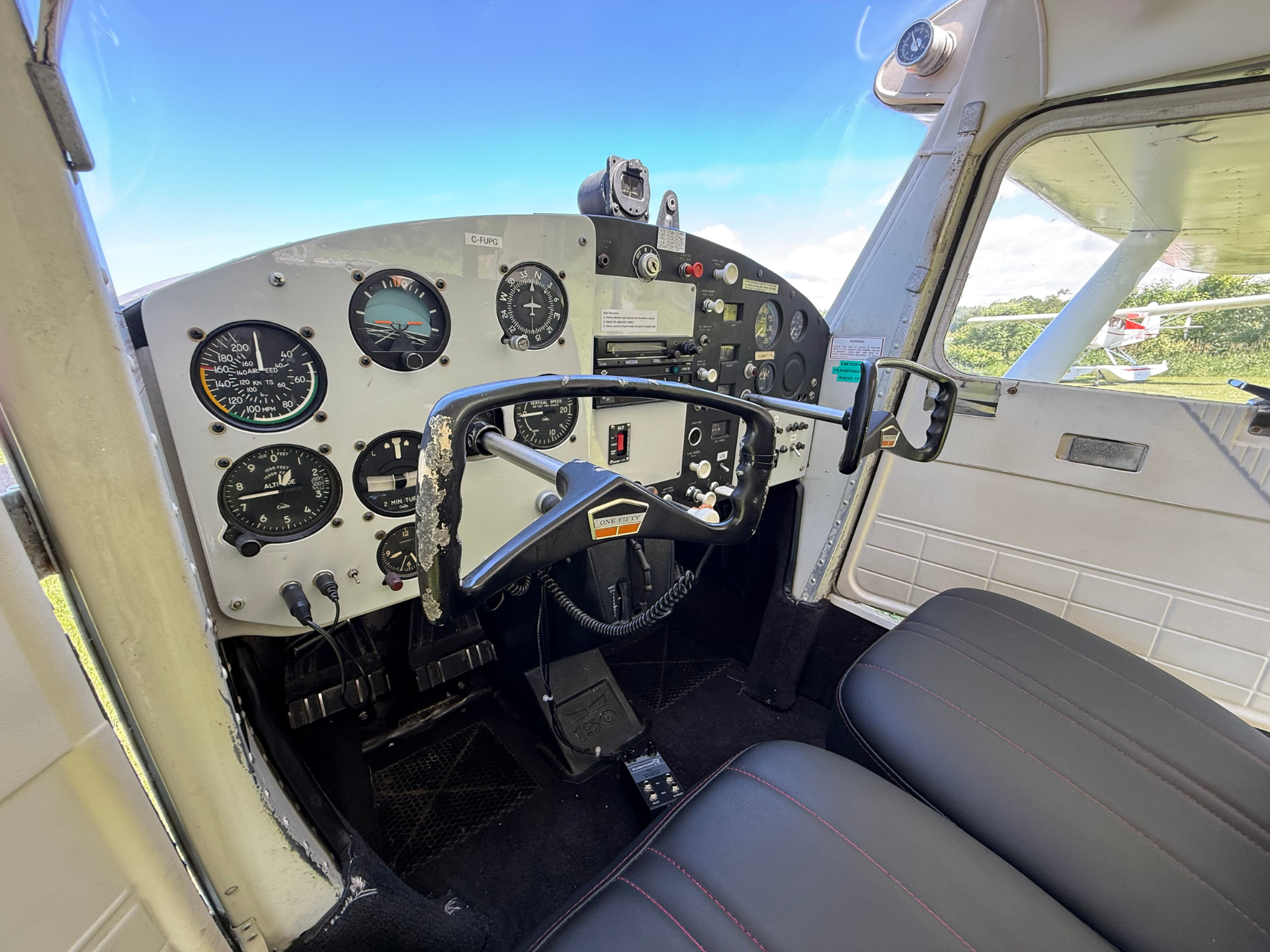 Interiors of a small airplane cockpit with analog gauges, yoke, and seats, view through open door to outside runway/airfield.