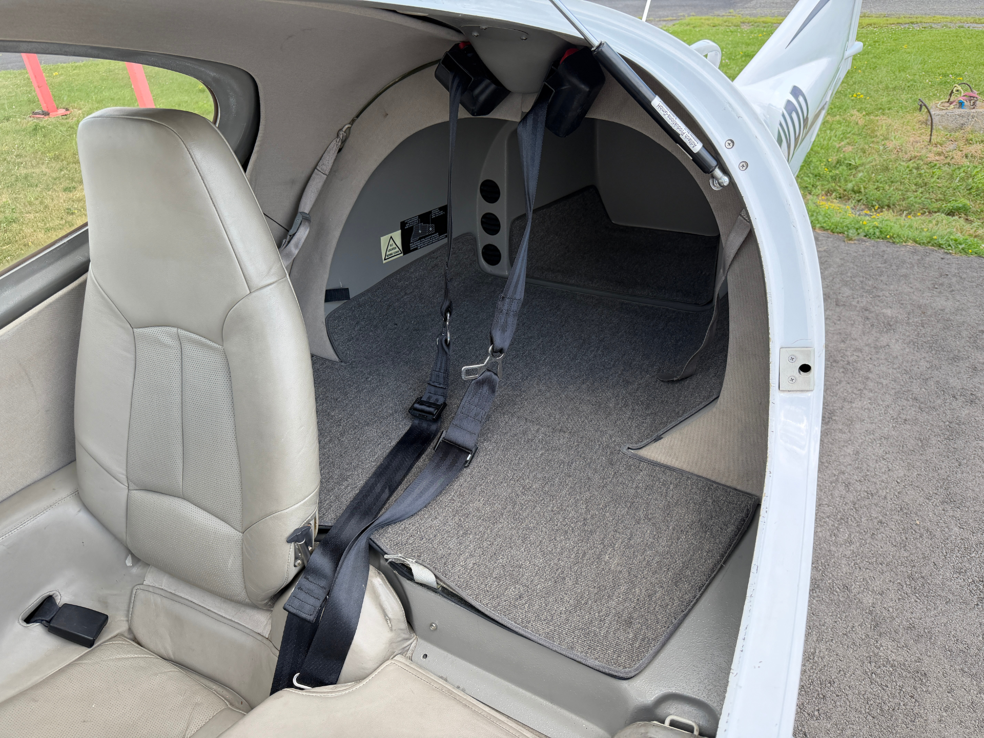 Small airplane cabin interior with beige seats and multiple black seat belts hanging, open door revealing gray carpet and cockpit area