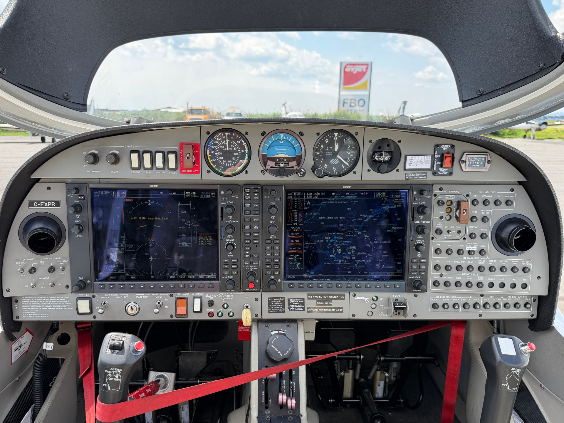 Cockpit of a small airplane showing multiple flight instruments, screens, switches, and red seat belts across the front panel.