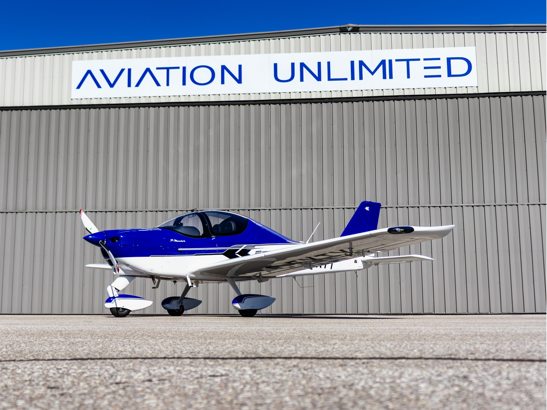 Blue and white small propeller plane on the tarmac in front of a gray hangar with an 'AVIATION UNLIMITED' sign above.