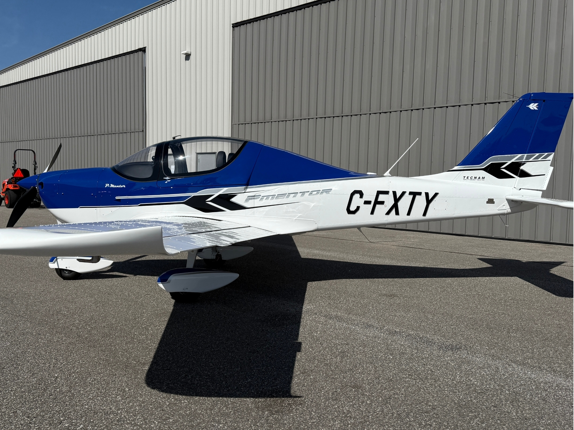 Blue and white small propeller airplane parked on a tarmac beside a gray metal hangar under a clear sky.
