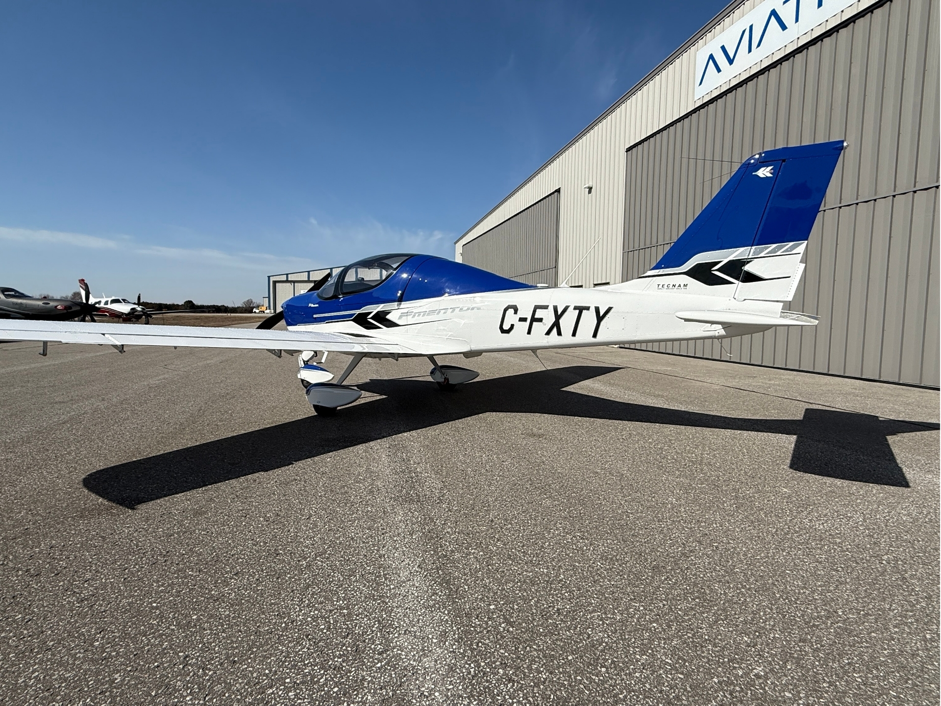 Blue-and-white small airplane with registration C-FXTY parked on a tarmac in front of a large hangar, clear blue sky overhead.