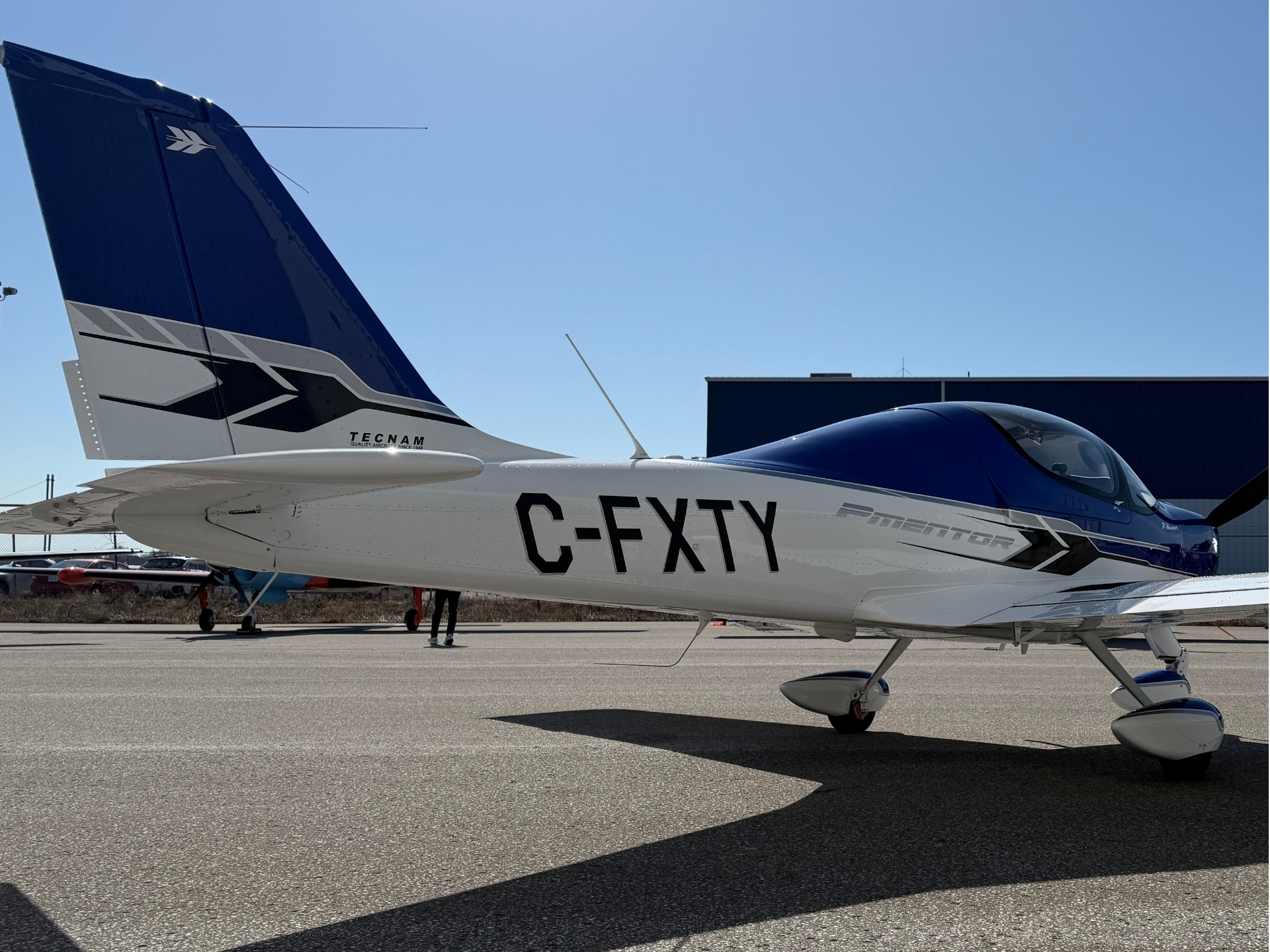 Small blue-and-white single-engine airplane on the tarmac, registration C-EXTY, Tecnam branding on tail.