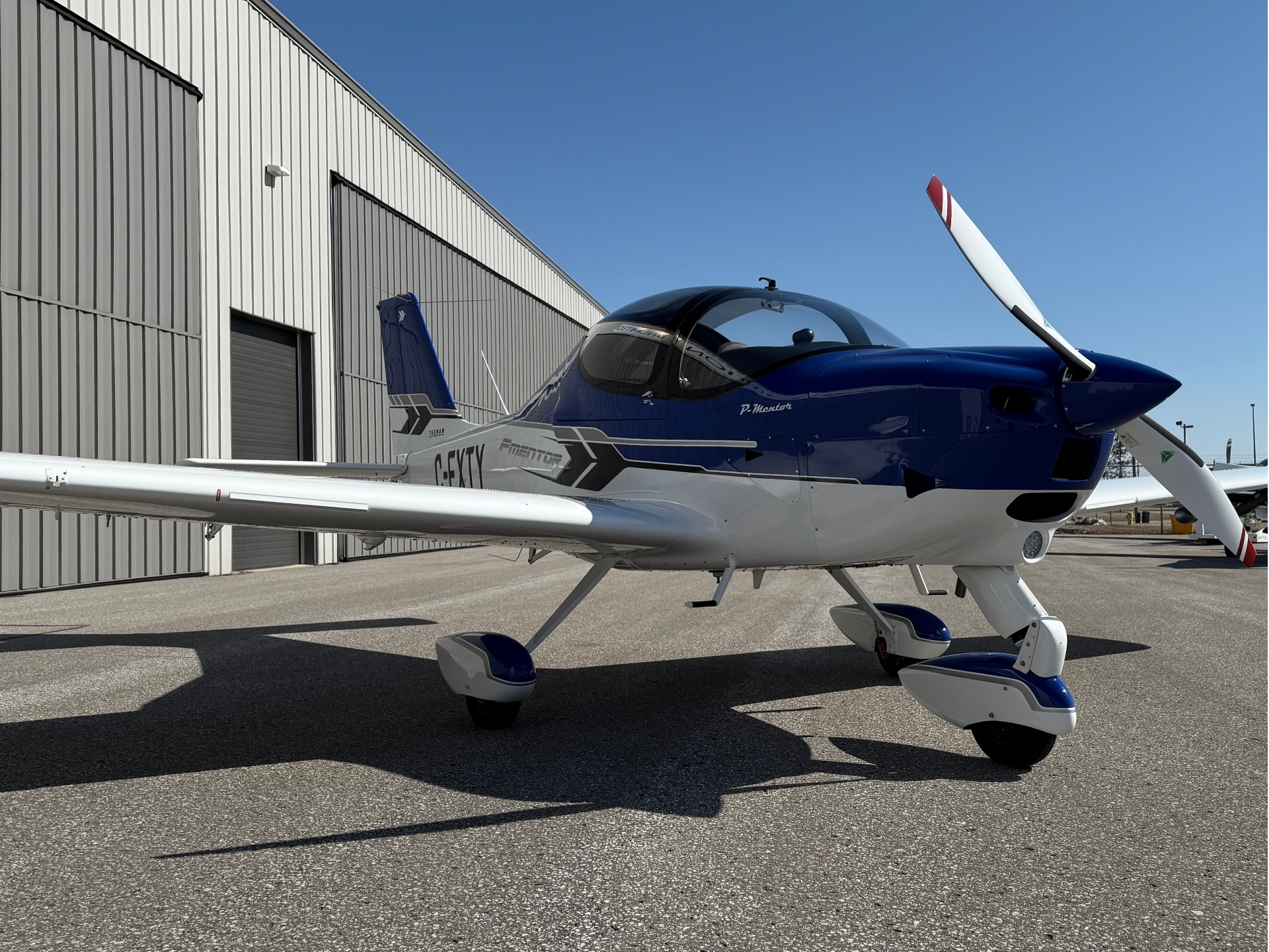 Blue and white single-engine propeller airplane parked on a tarmac beside a metal hangar.