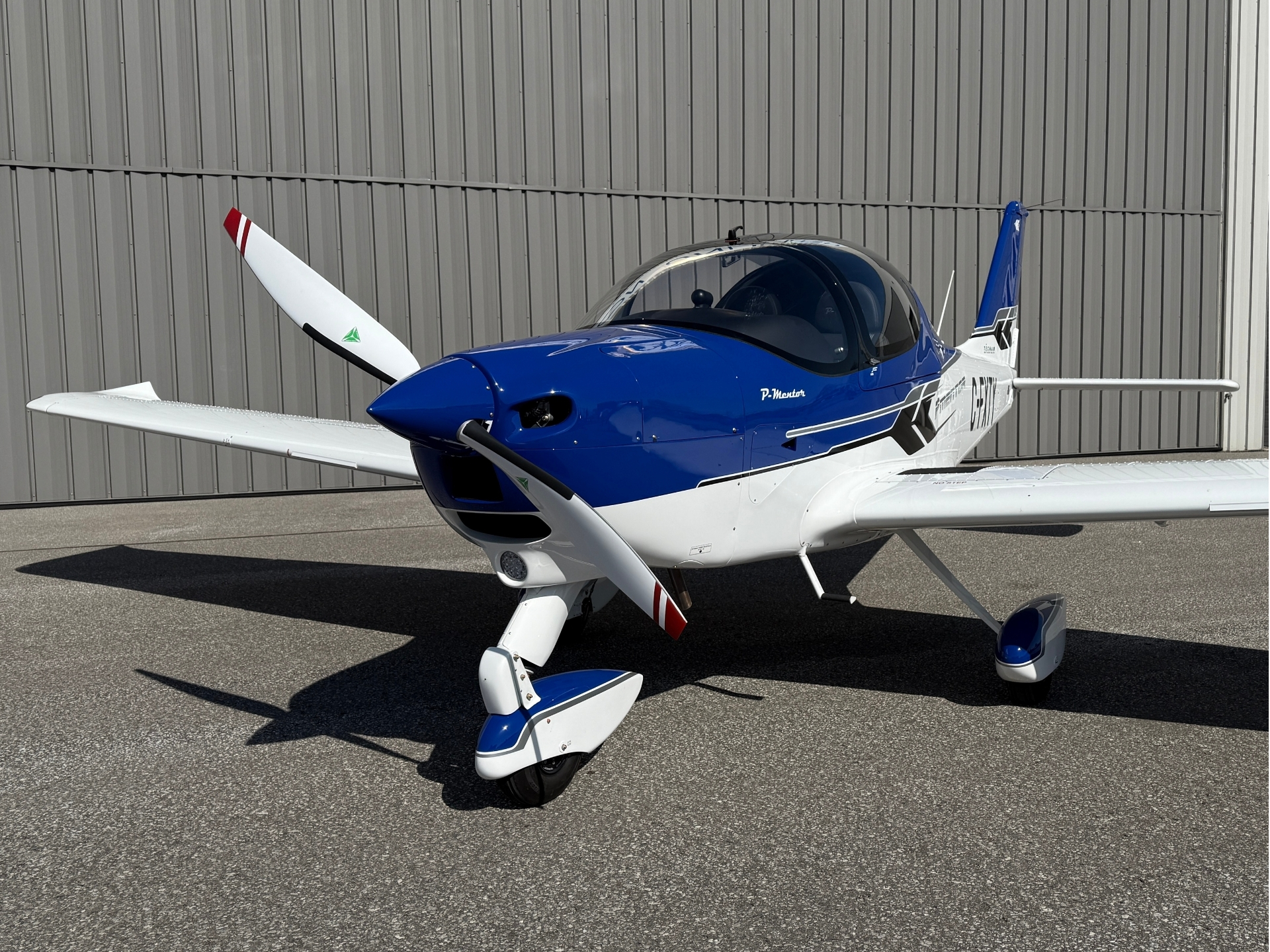 Blue and white single-engine propeller airplane parked on the tarmac in front of a gray hangar, side view of cockpit and nose gear visible