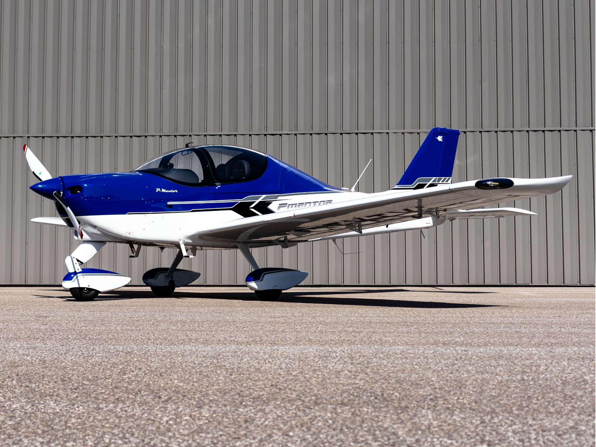 Blue and white single-engine airplane parked on the tarmac in front of a gray hangar wall.