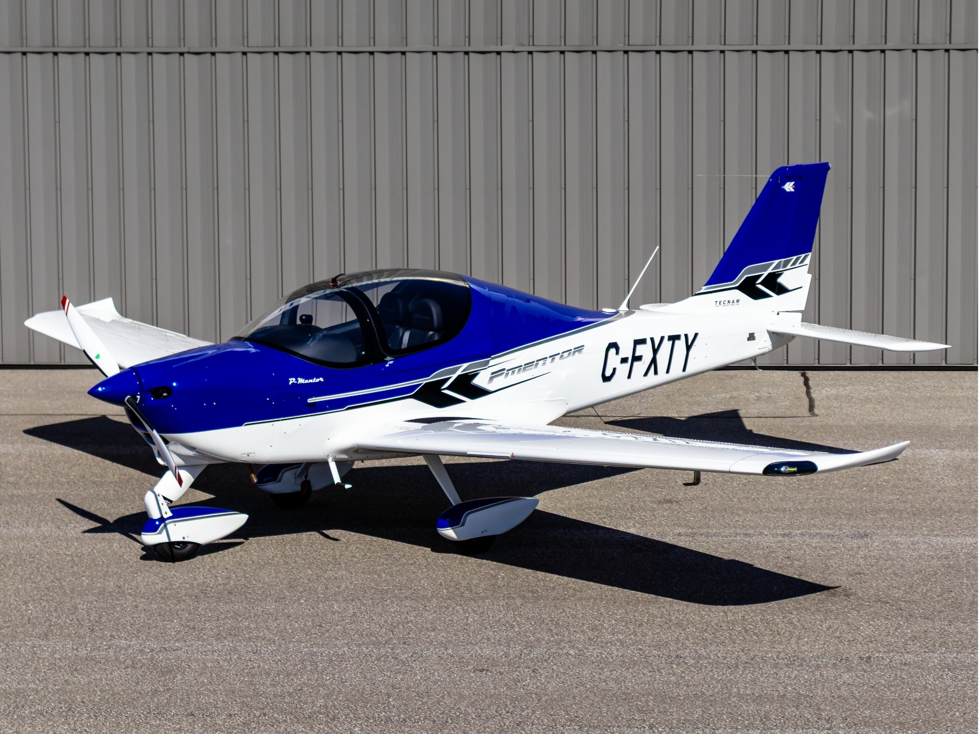 Small blue-and-white light aircraft parked on tarmac with a grey hangar in the background; registration C-FXTY visible on fuselage