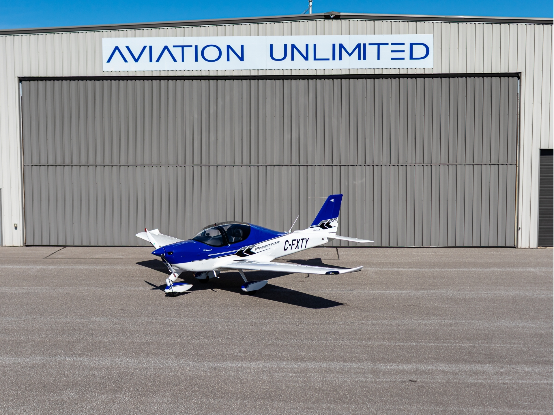 Blue and white small aircraft on the tarmac in front of a gray hangar with 'Aviation Unlimited' sign above the door
