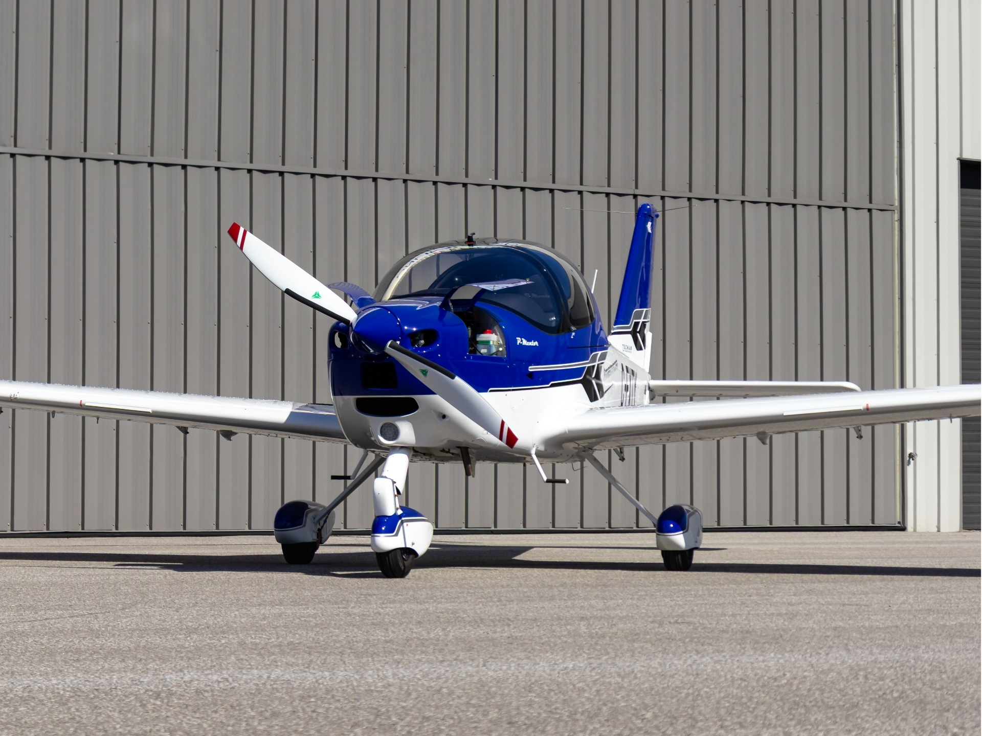 Blue and white small propeller airplane parked on a tarmac with a gray hangar backdrop in the background; cockpit canopy visible in blue/gray hues.