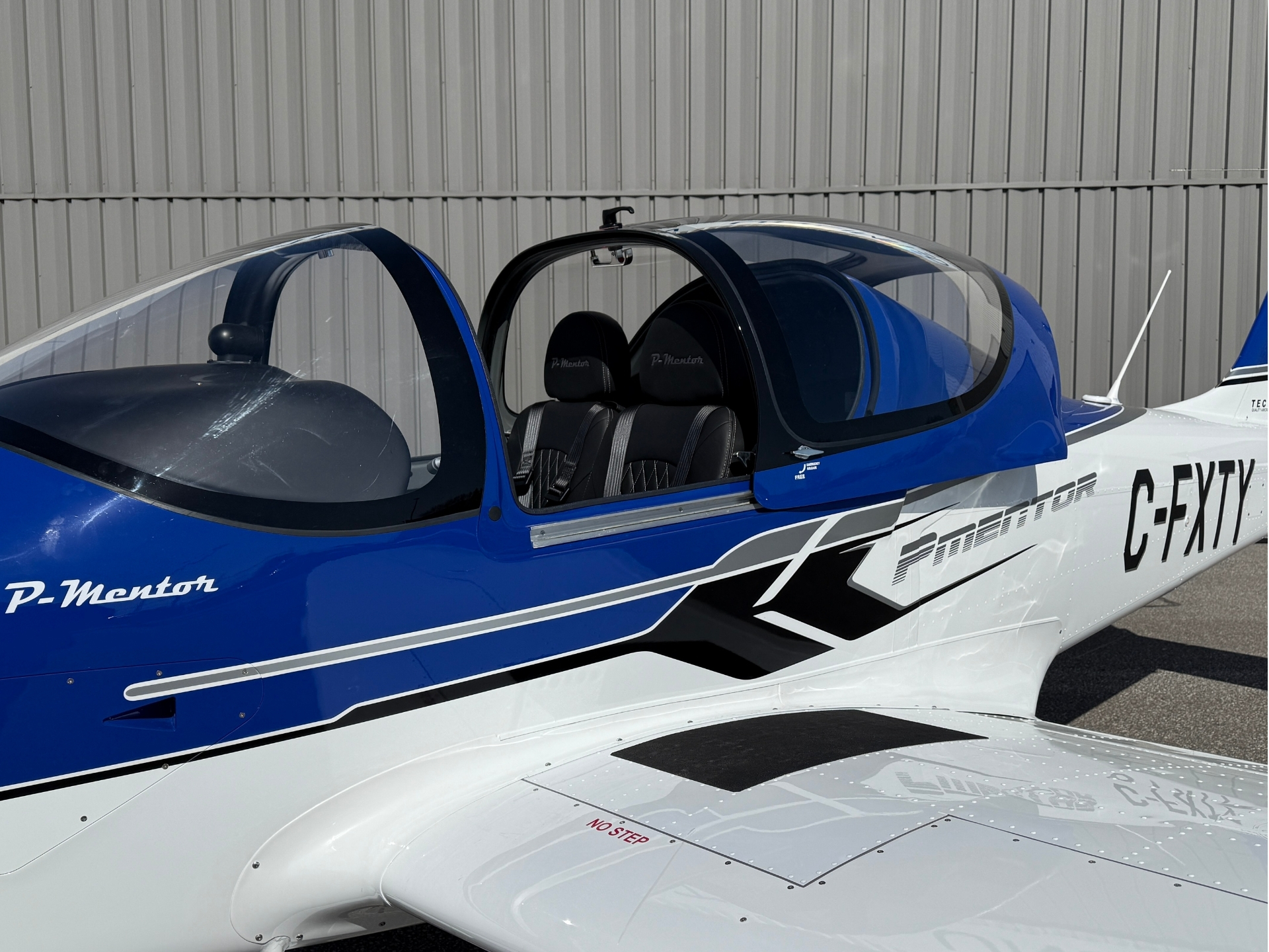 Blue and white light aircraft with an open cockpit and two black seats, parked beside a gray hangar wall.