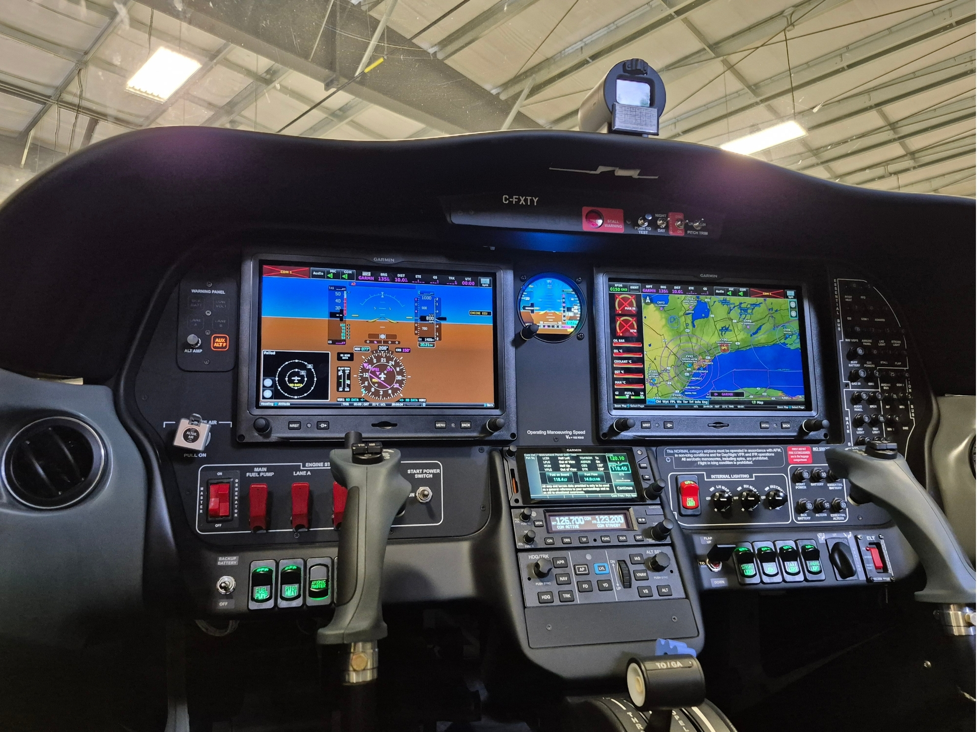 Glass cockpit of a small aircraft with two Garmin displays, map screen, and flight controls visible in the foreground, over a hangar interior.