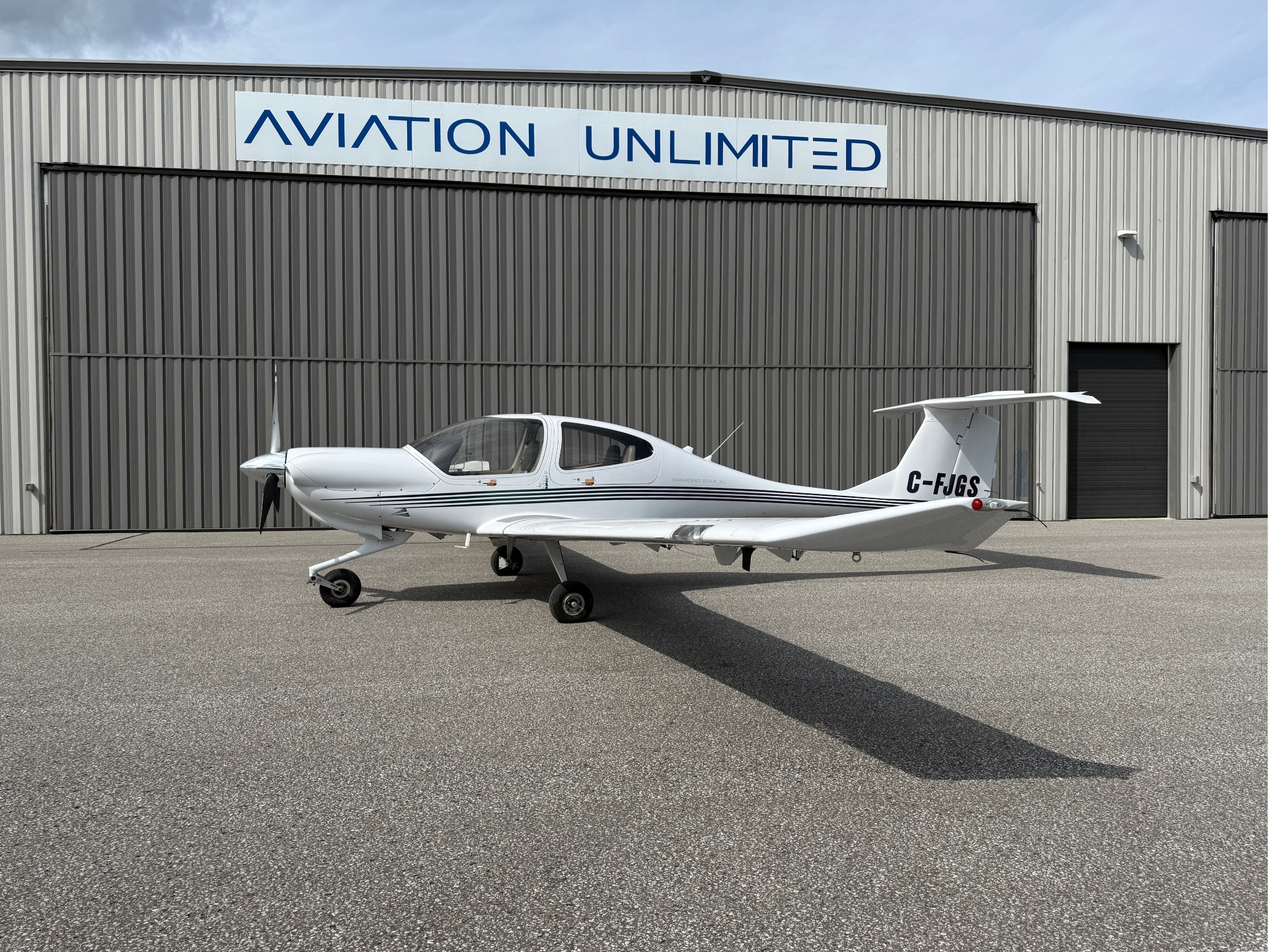 White single-engine propeller plane parked on a tarmac in front of a gray metal hangar with an Aviation Unlimited sign above it.
