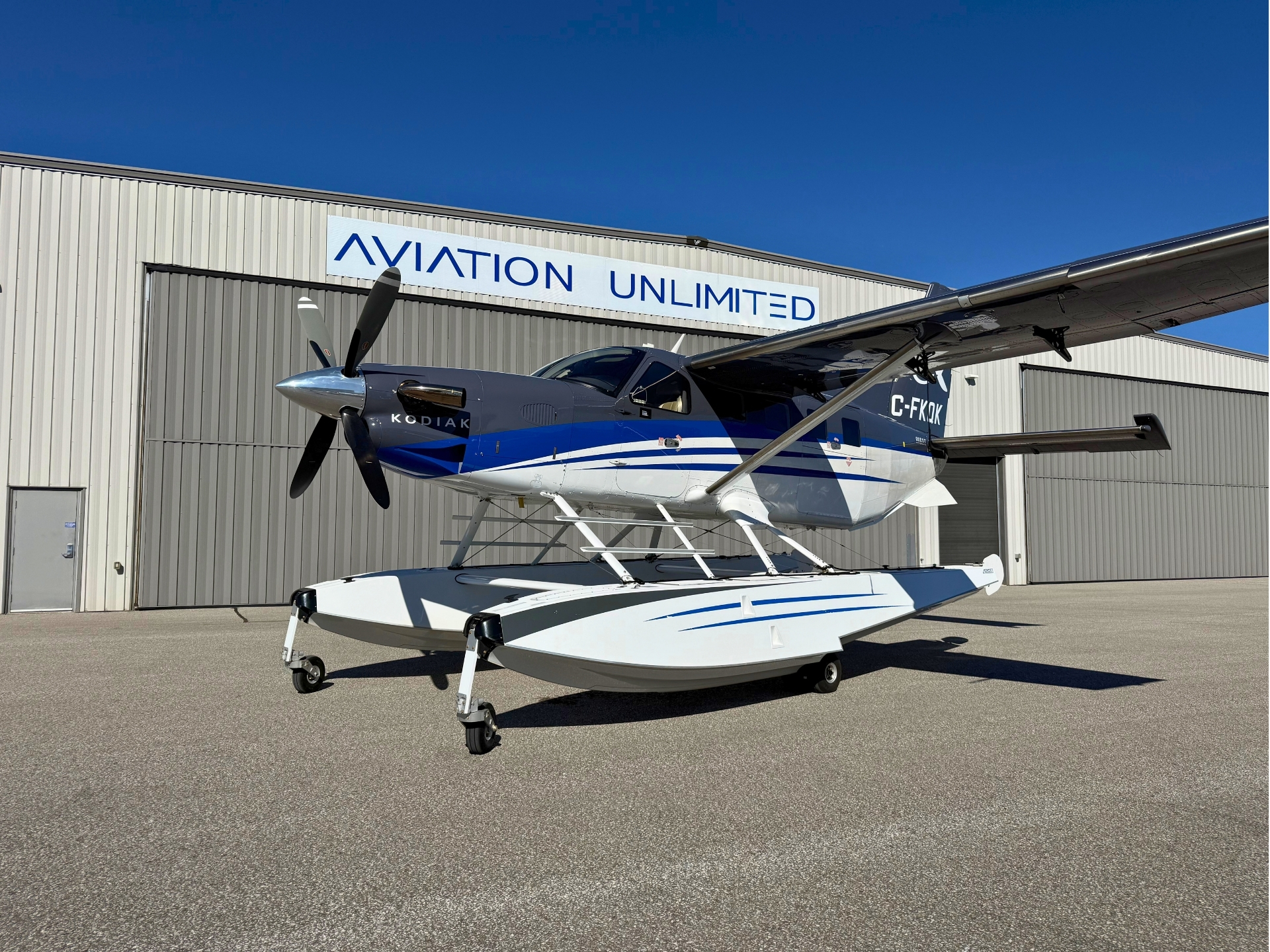 Blue and white Kodiak floatplane on its landing gear parked in front of a hangar reading AVIATION UNLIMITED under a clear blue sky