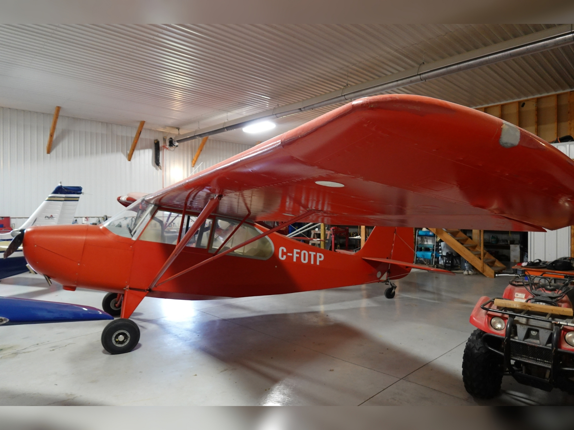 Red single engine airplane in a hangar with another aircraft and maintenance gear visible in the background