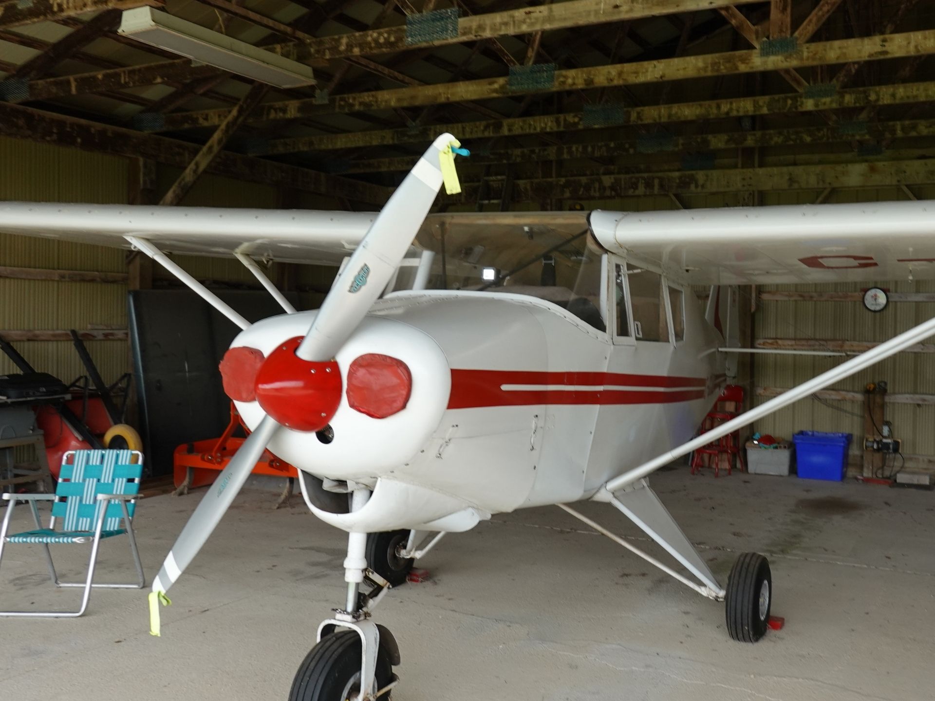 Small single-engine airplane with red nose and stripe parked inside a hangar with wooden beams overhead and equipment around it.