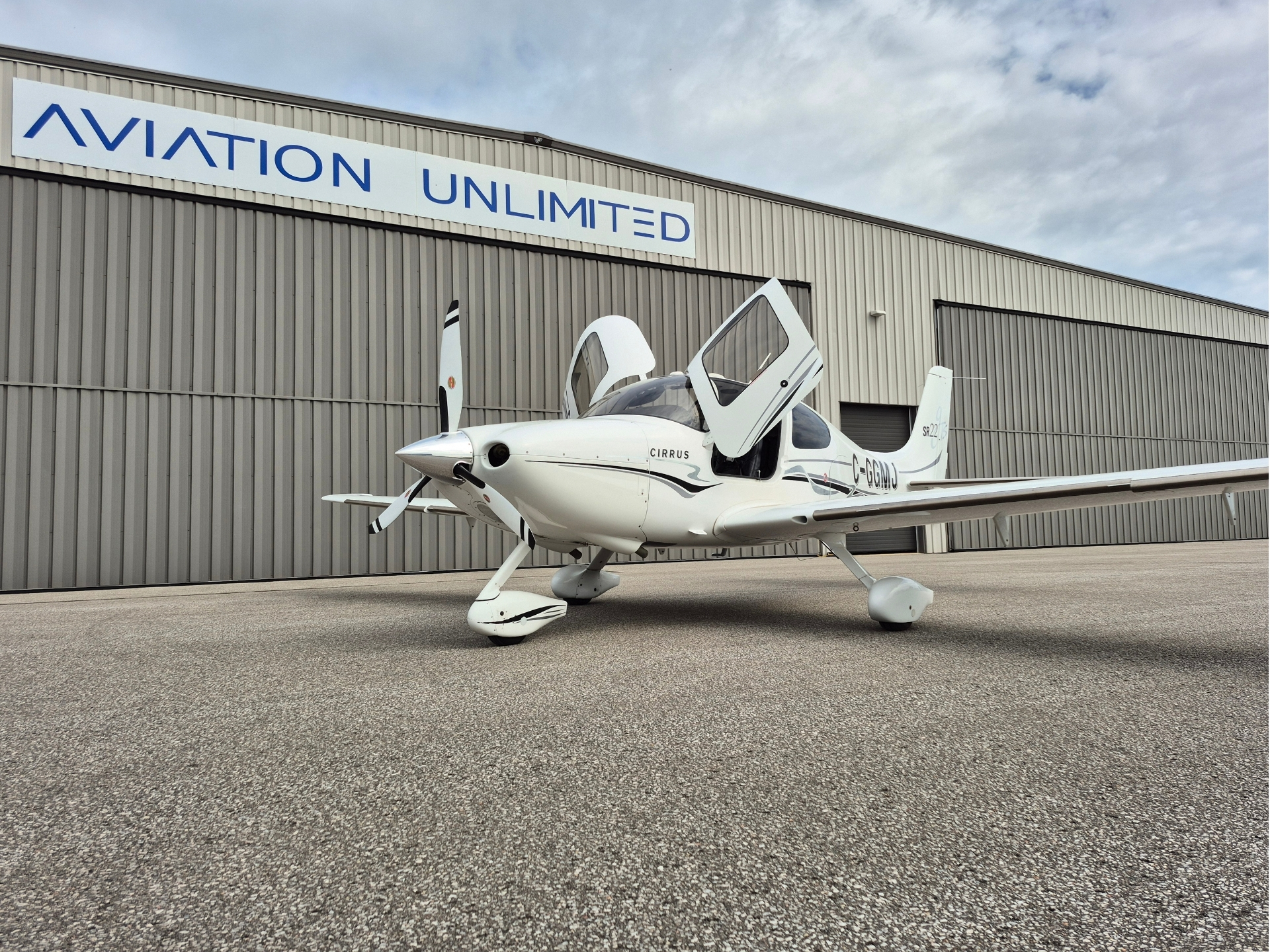 White Cirrus single engine airplane with open doors on a tarmac in front of a gray hangar reading AVIATION UNLIMITED