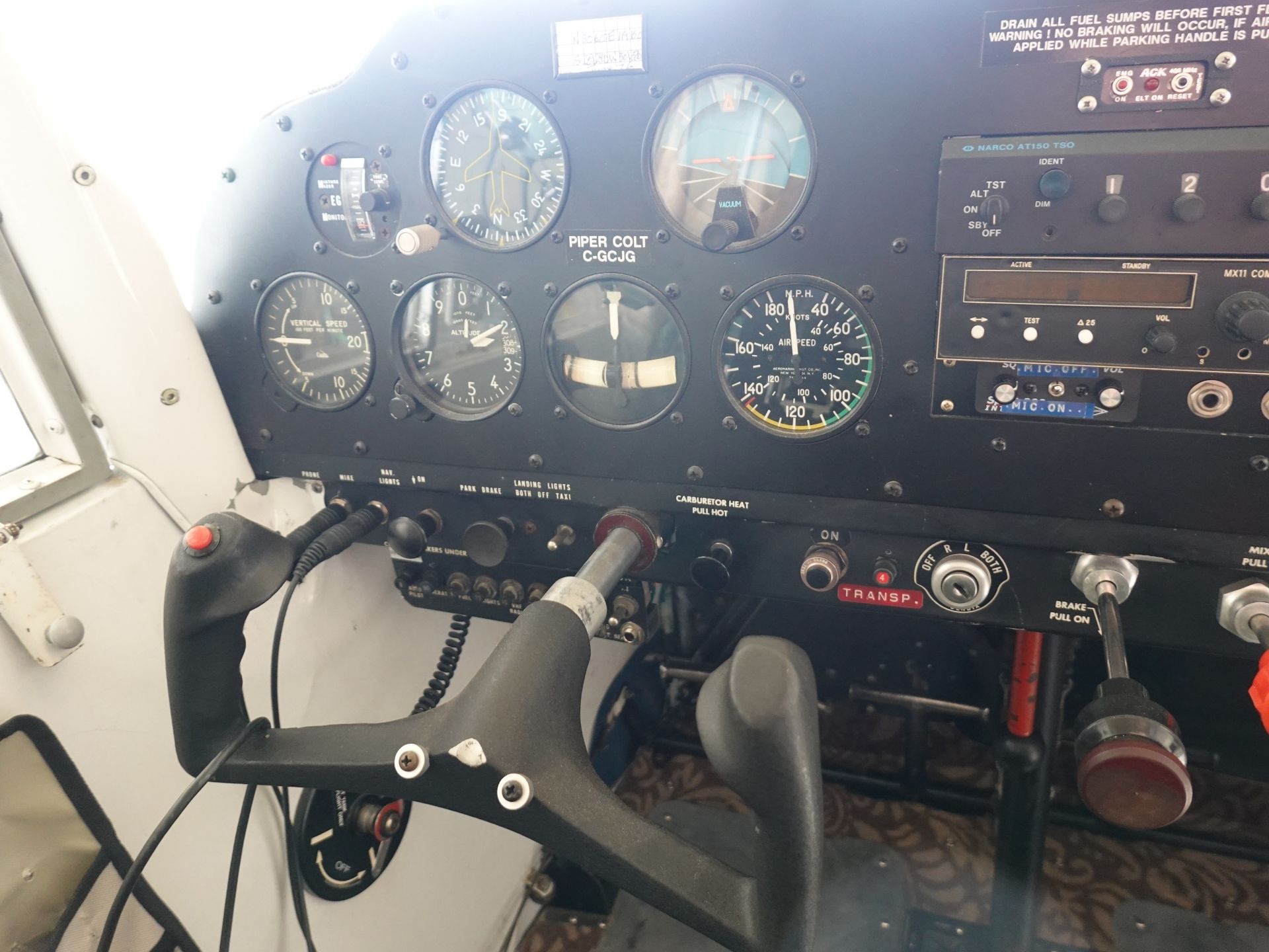 Cockpit of small airplane with multiple gauges, yoke, and control panel labeled Piper Colt C-GCJG visible behind the instrument cluster.